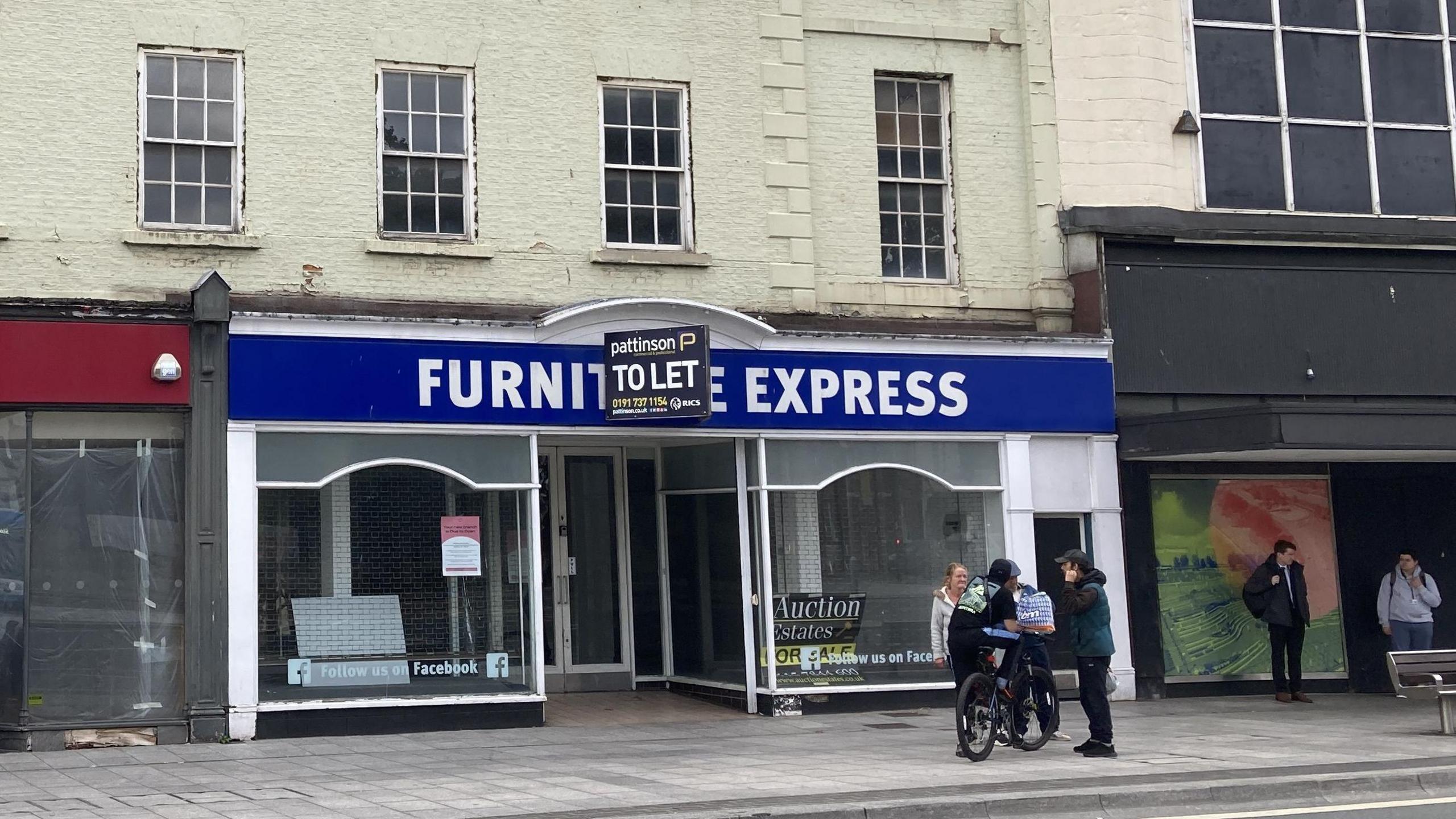 The former Furniture Express store in Stockton. A "To Let" sign is fixed above the doorway. There are large glass windows to either side. A small number of people are chatting on the pavement in front of the building. The upper floors are painted cream.