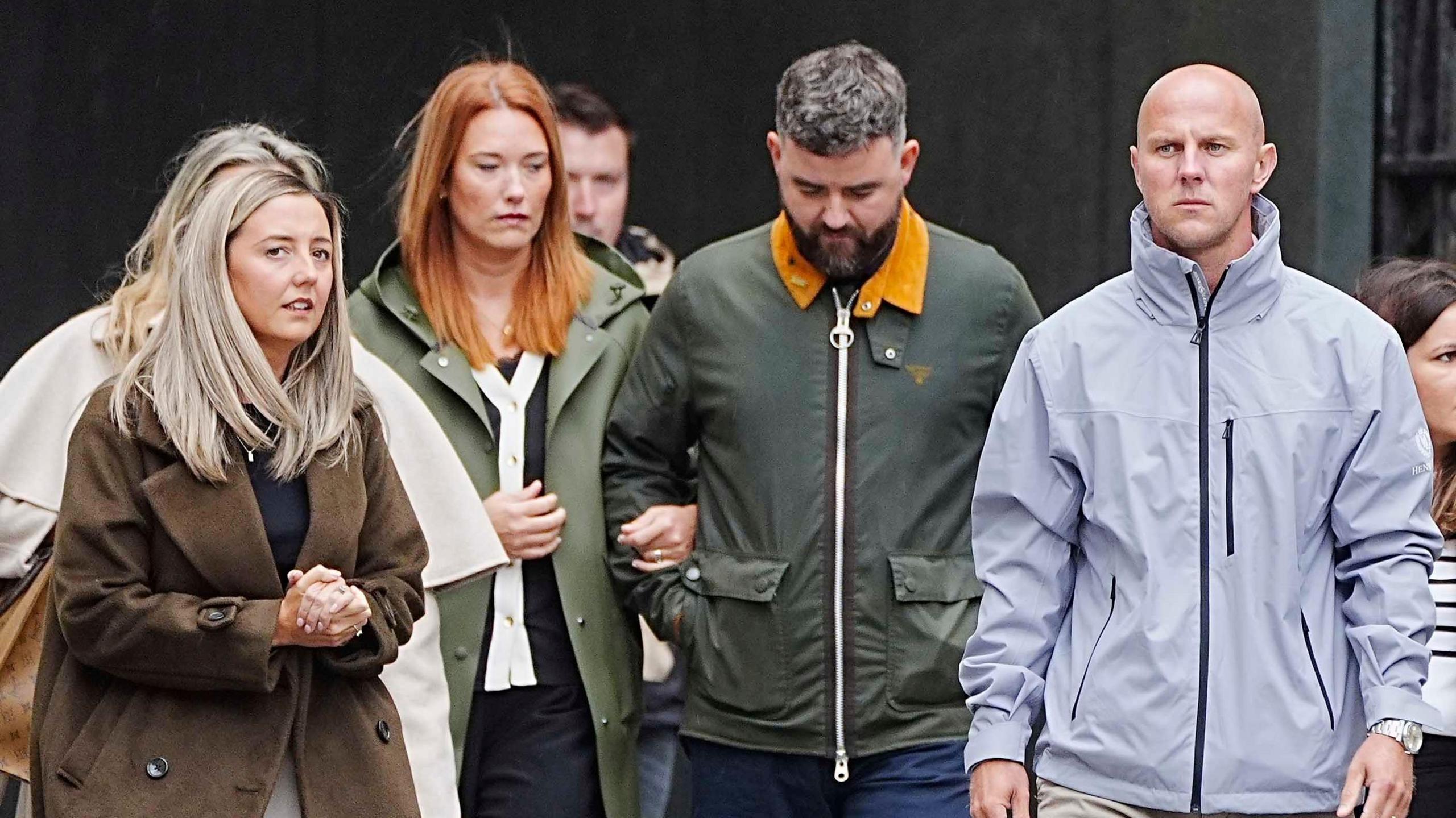 Jenni Stancombe, with long blonde hair and a brown coat, walks with her hands clasped together. David Stancombe, who has a bald head and is wearing a grey coat, walks a few paces from her.