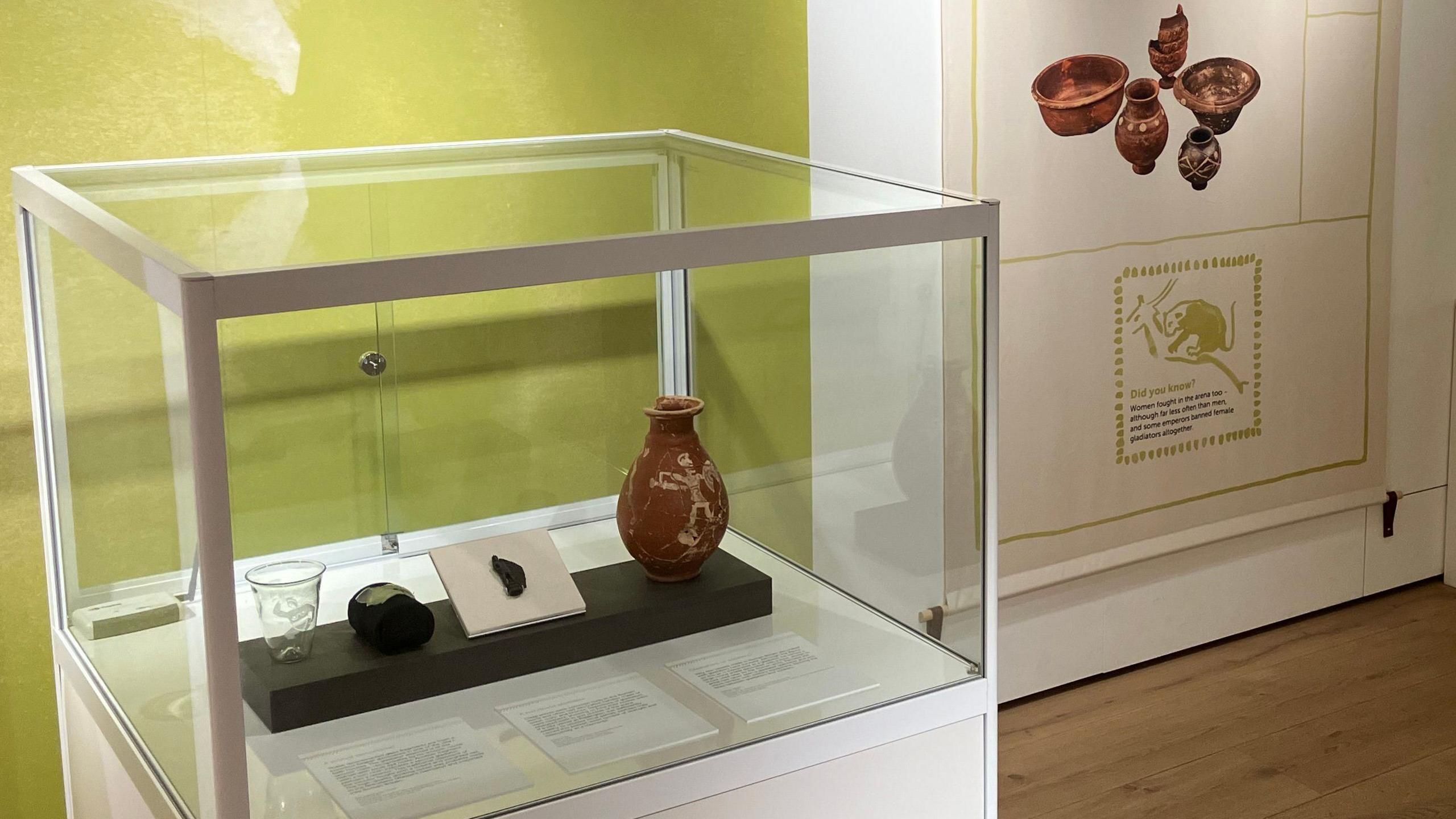 A glass cabinet containing a glass cup, two small urns and a shard of glass.