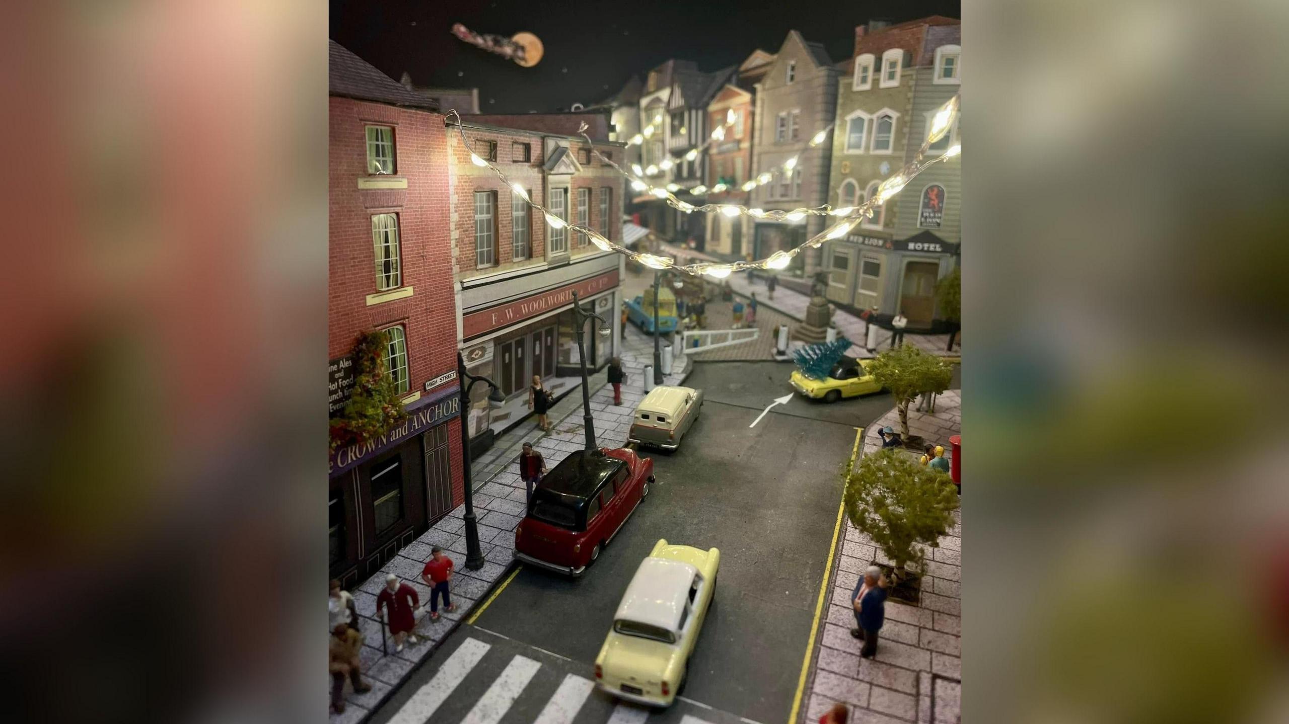 A model village which shows vintage cars driving through a recreation in miniature of the bottom of Guildford High Street, complete with barrier stopping cars going up the cobbled part of the high street. There are fairy lights strung between the buildings and model people walking on the pavements.