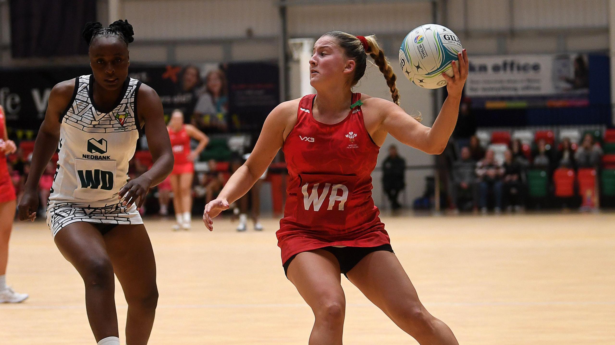 Bethan Dyke in the red of Wales holds the ball high in her left hand alongside a Zimbabwe player in a black and white dress