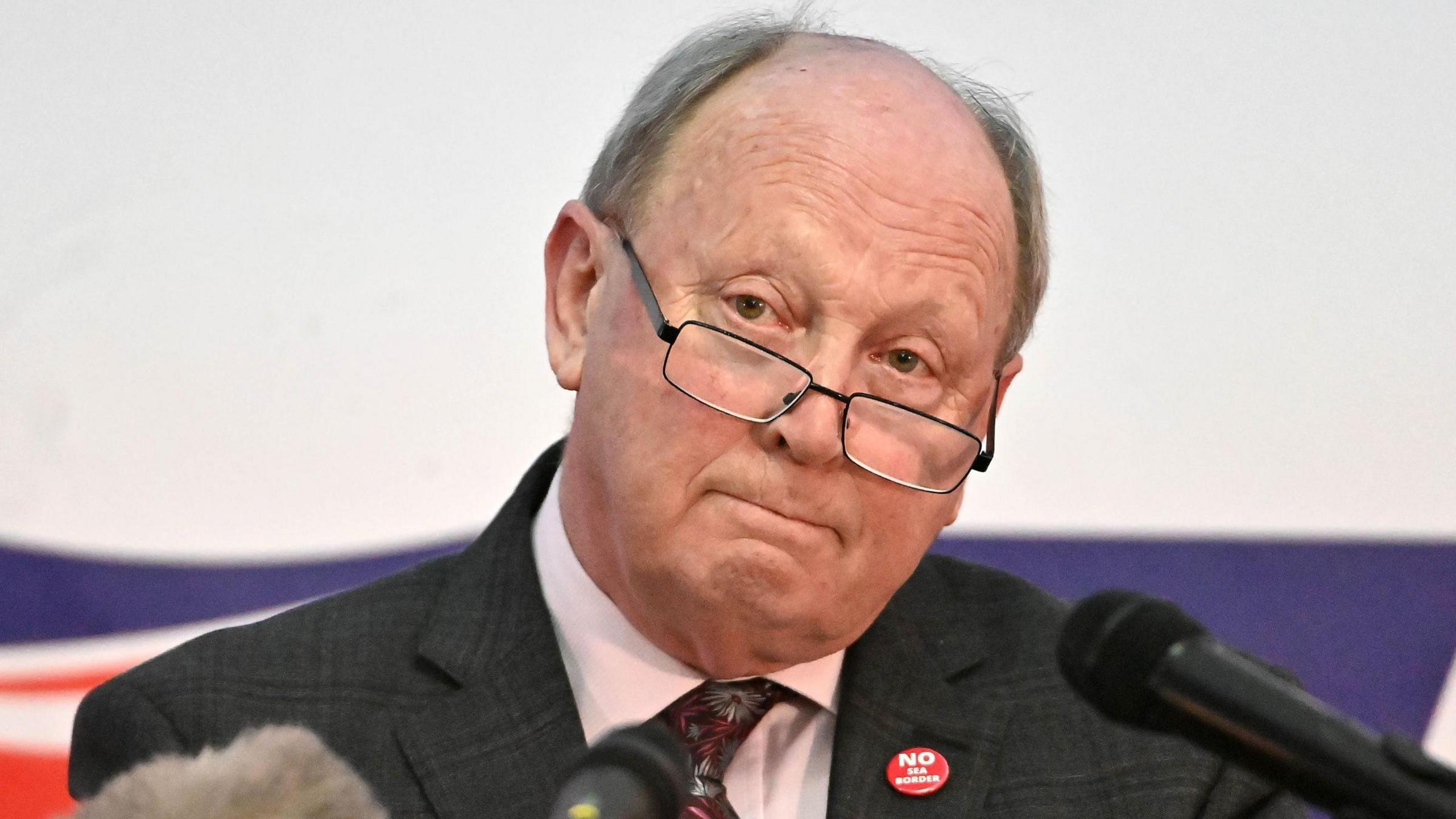 A man is standing in front of news microphones. He is wearing a dark suit, white shirt and tie. He has glasses on and is bald with greying hair to the sides.