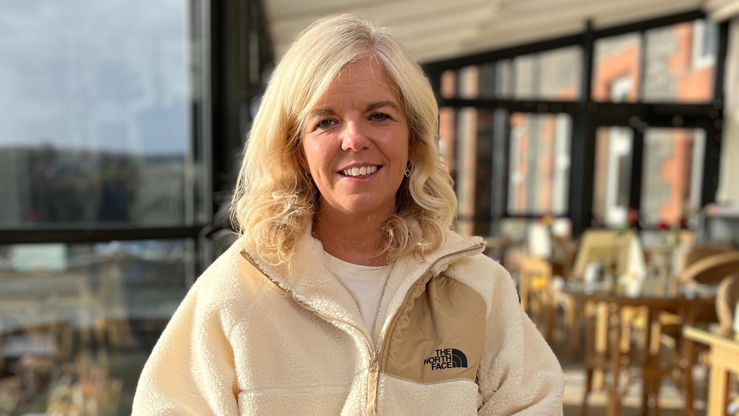 A woman with blonde-curly hair is wearing a cream borg material jacket with branding on it. 