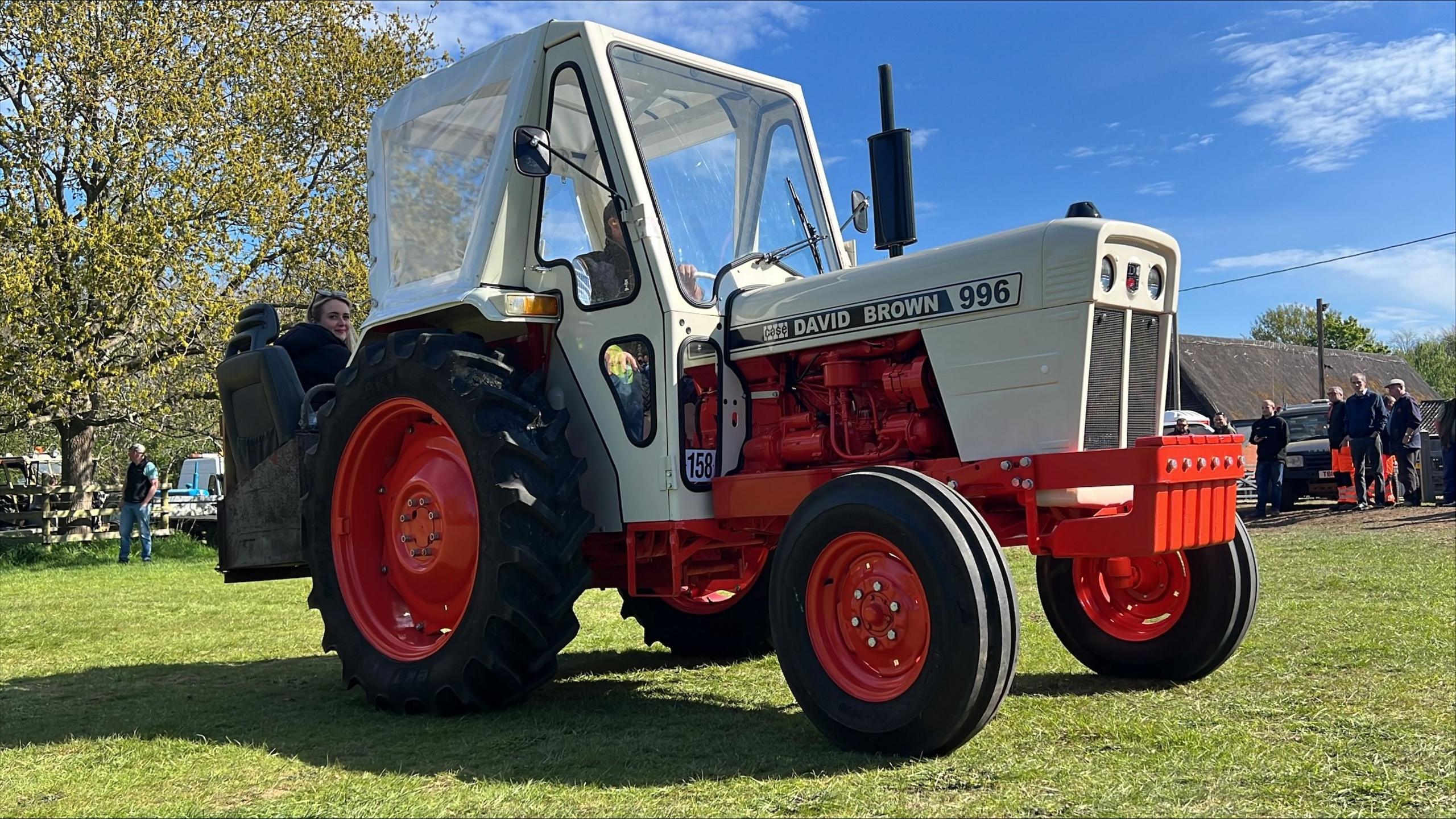 A vintage white and red David Brown tractor parked in a field. It has large back wheels and smaller front ones. It has a spacious cabin with big glass windows.