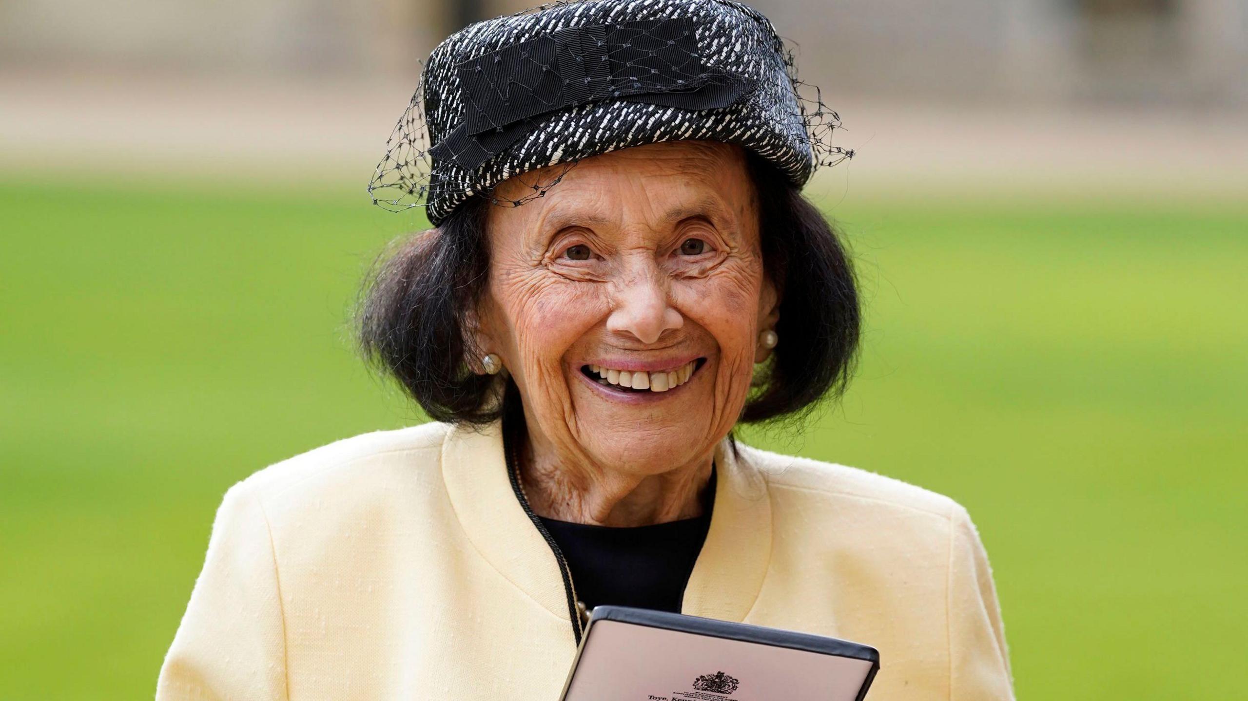 Lily after being made a MBE at Windsor Castle in January 2023. She has short, black hair and is smiling. She is wearing a cream jacket and black and silver hat.