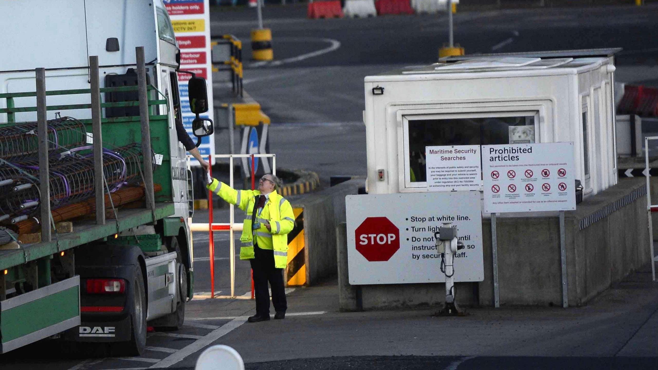 A man wearing a yellow yellow high-viz jacket is checking documents being handed to him by the driver of a lorry. There is a cabin in the backgound and signs giving information about prohibited items.