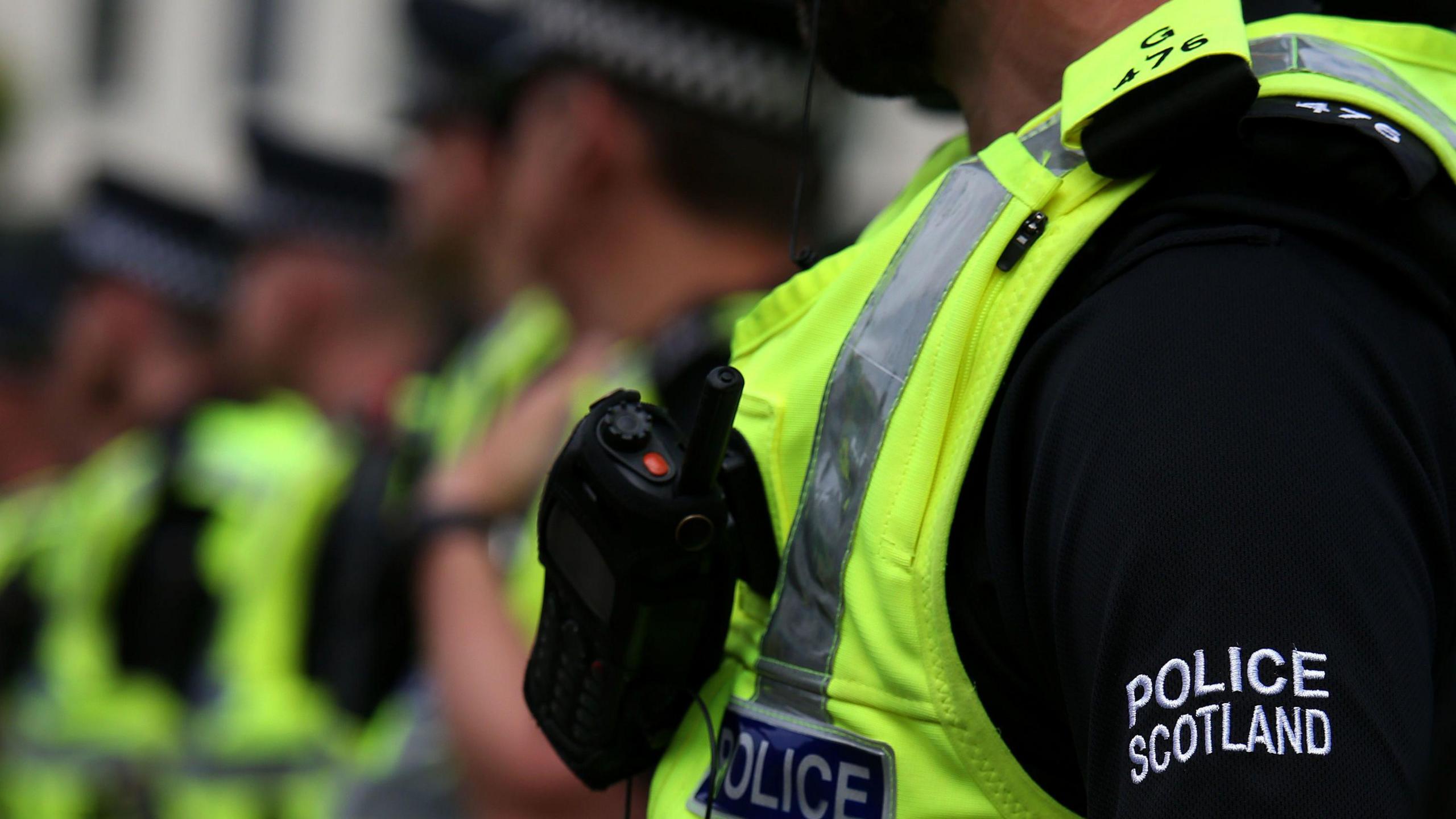 A generic photo of a row of Police Scotland officers, their faces blurred and the focus on the nearest officer, their radio and hi-vis gilet.