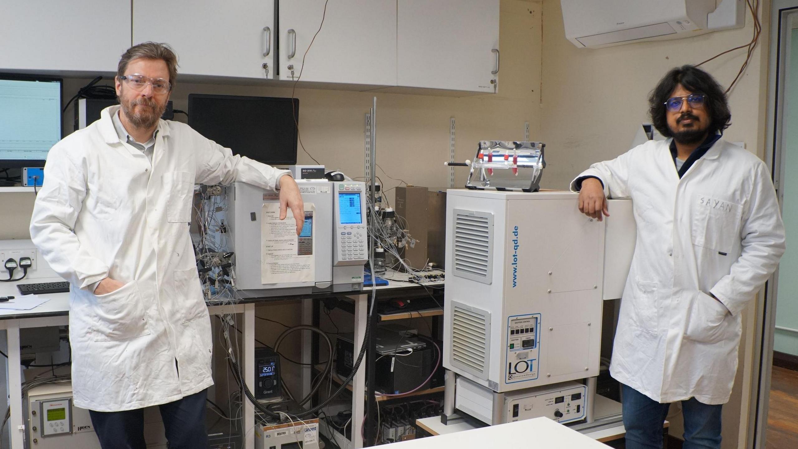 scientists Erwin Reiser (left) and Sayan Kar (right) in white lab coats standing either side of lots of machinery