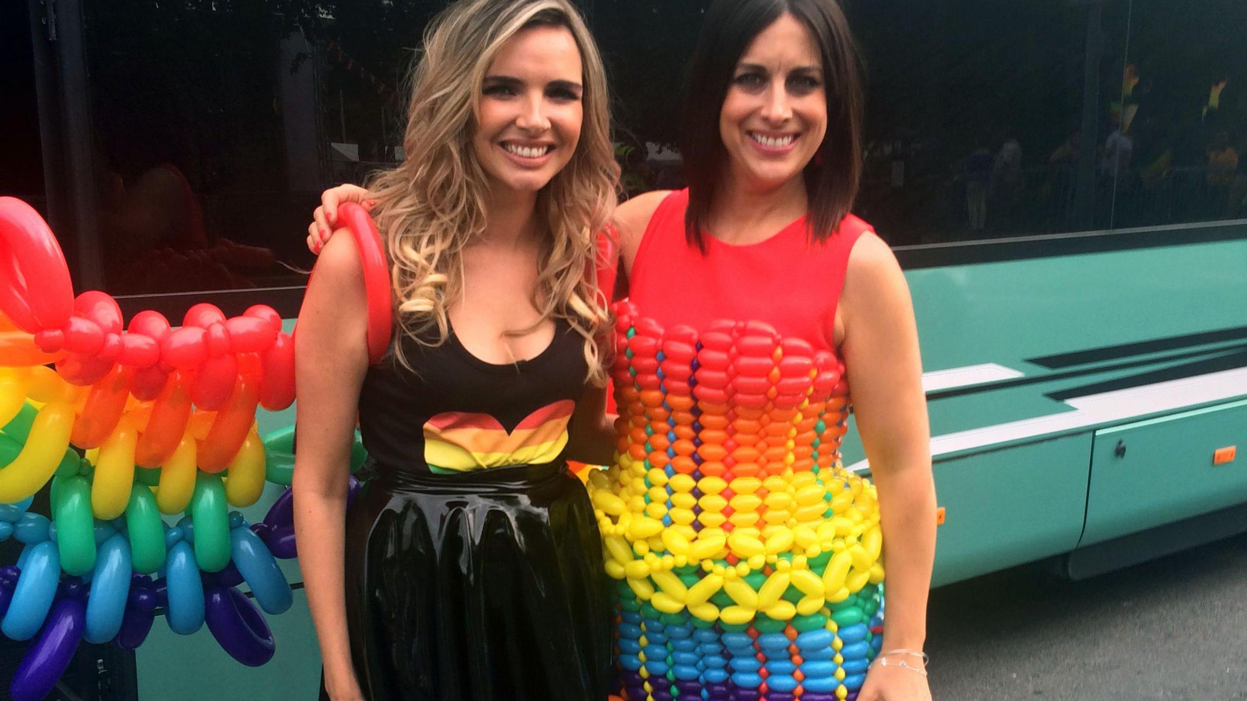 Pop star Nadine Coyle poses with Irish TV presenter Lucy Kennedy, who has her arm around Nadine. Both are smiling at the camera and standing in front of a light green coach. Nadine is wearing a black shiny skirt and a vest with a rainbow heart on the front. She is wearing a set of wings made out of balloons in rainbow colours. Lucy is wearing a red vest and a full balloon dress in rainbow colours.