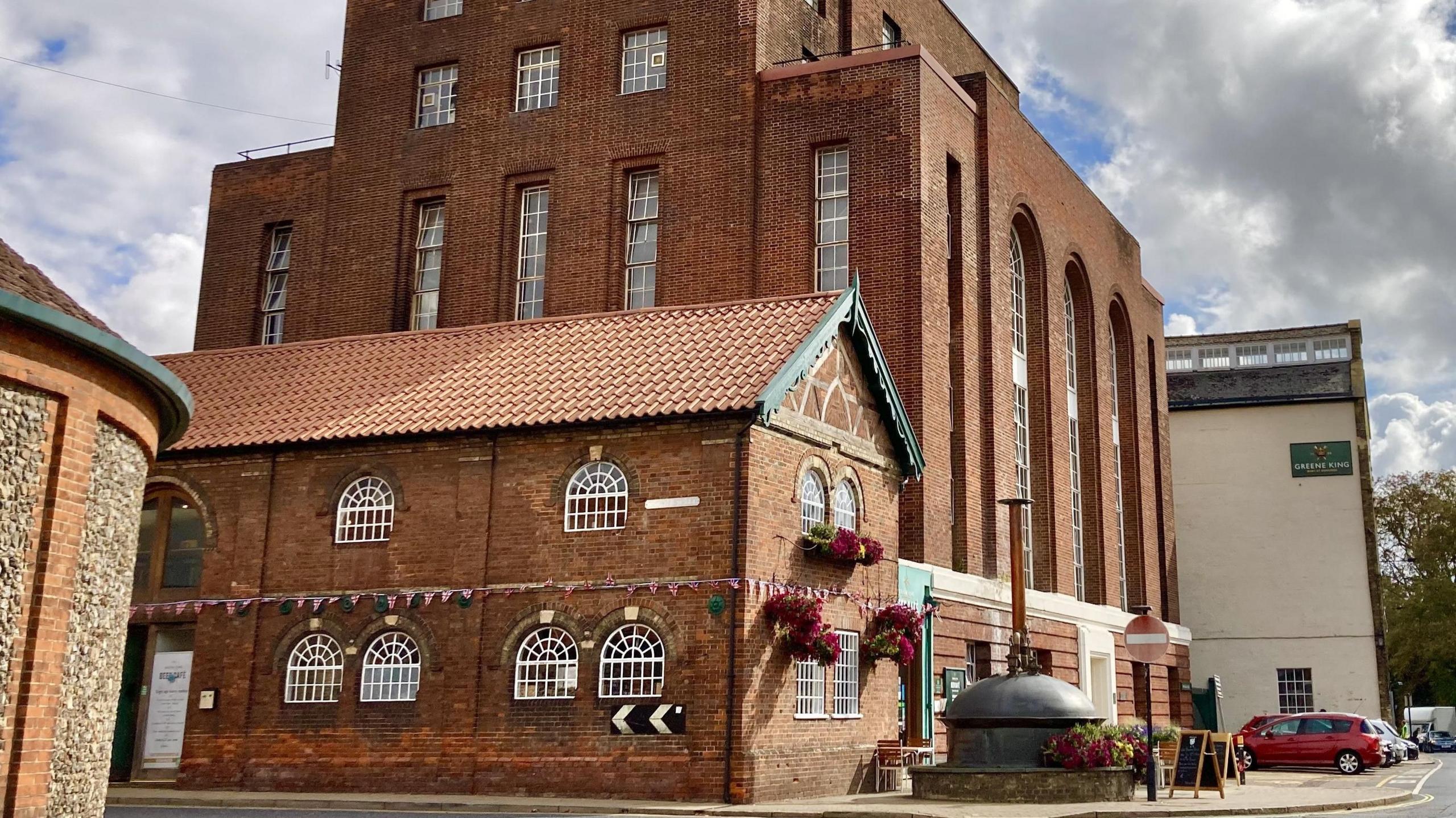 Greene King's brewery in Bury St Edmunds is pictured. It is a multi-storey red brick building with large windows. Cars can be seen parked up outside the building.