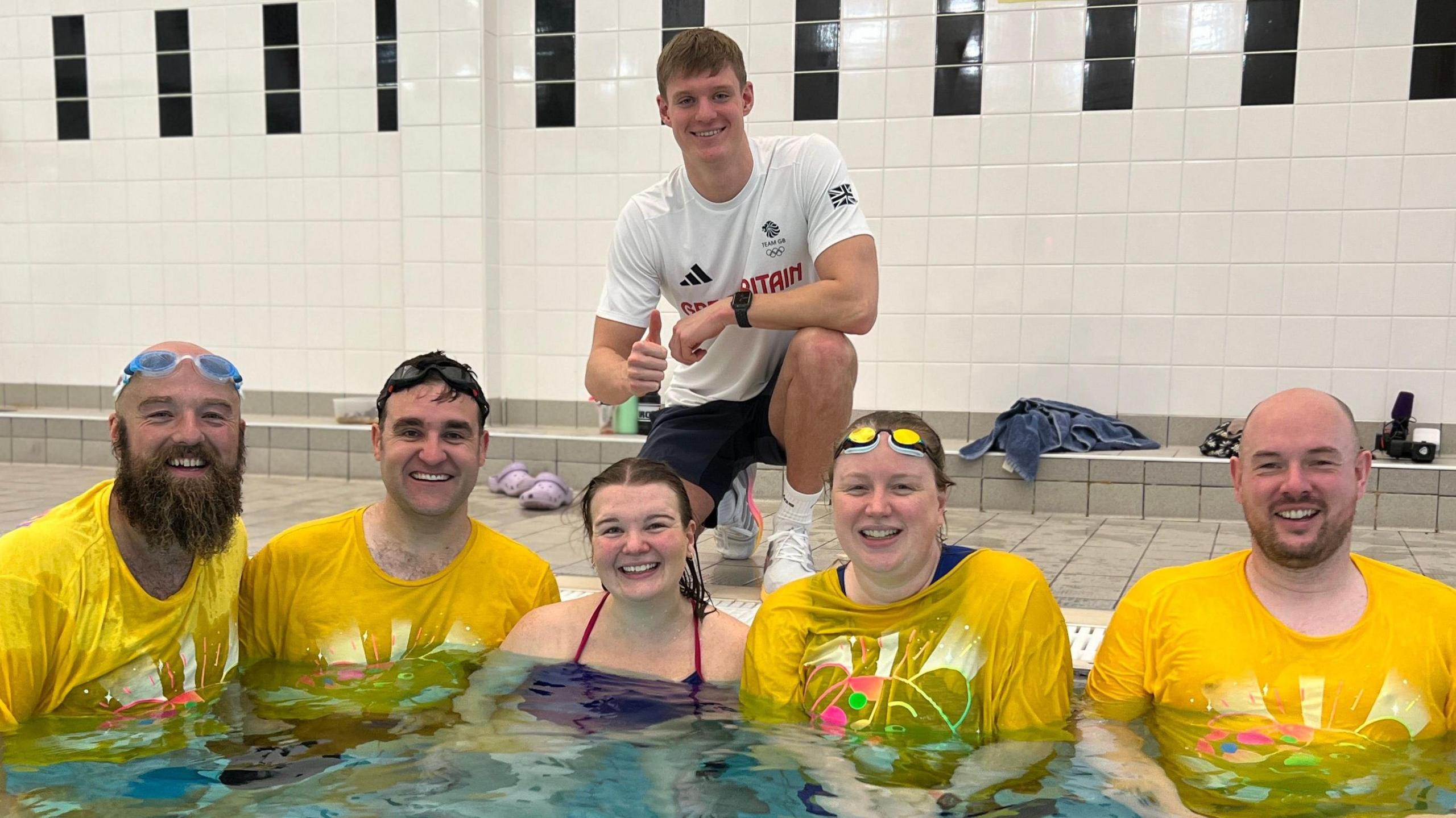 Oliver Morgan is crouching on the side of a swimming pool wearing a white T-shirt. Below him in the pool are five people. Two men on the left are wearing yellow Children in Need T-shirts, a woman is wearing a swimming costume. To her right is a woman and a man, both also wearing yellow Children in Need T-shirts