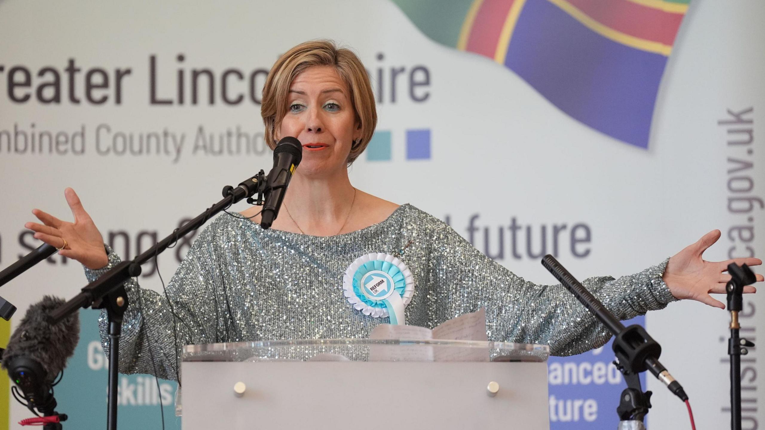 Dame Andrea Jenkyns stands at a podium surrounded by four black microphones. She has her arms open and is wearing a sparkly silver dress with a blue and white rosette.