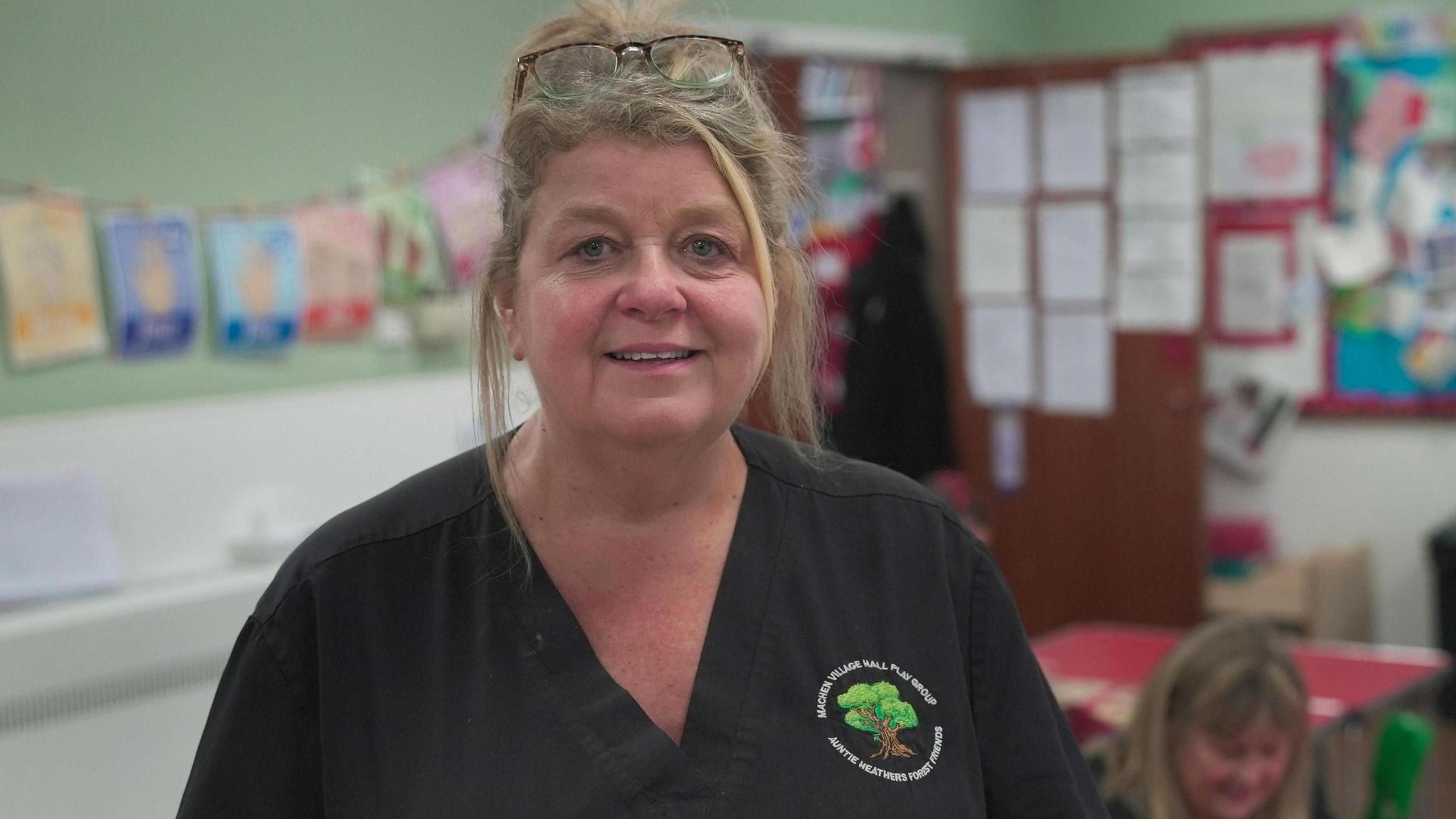 Heather Street - A woman with a black shirt on with the logo of a tree and the name of the nursery in a white font. She looks at the camera and has her blonde hair tied up. It is a head and shoulders shot.