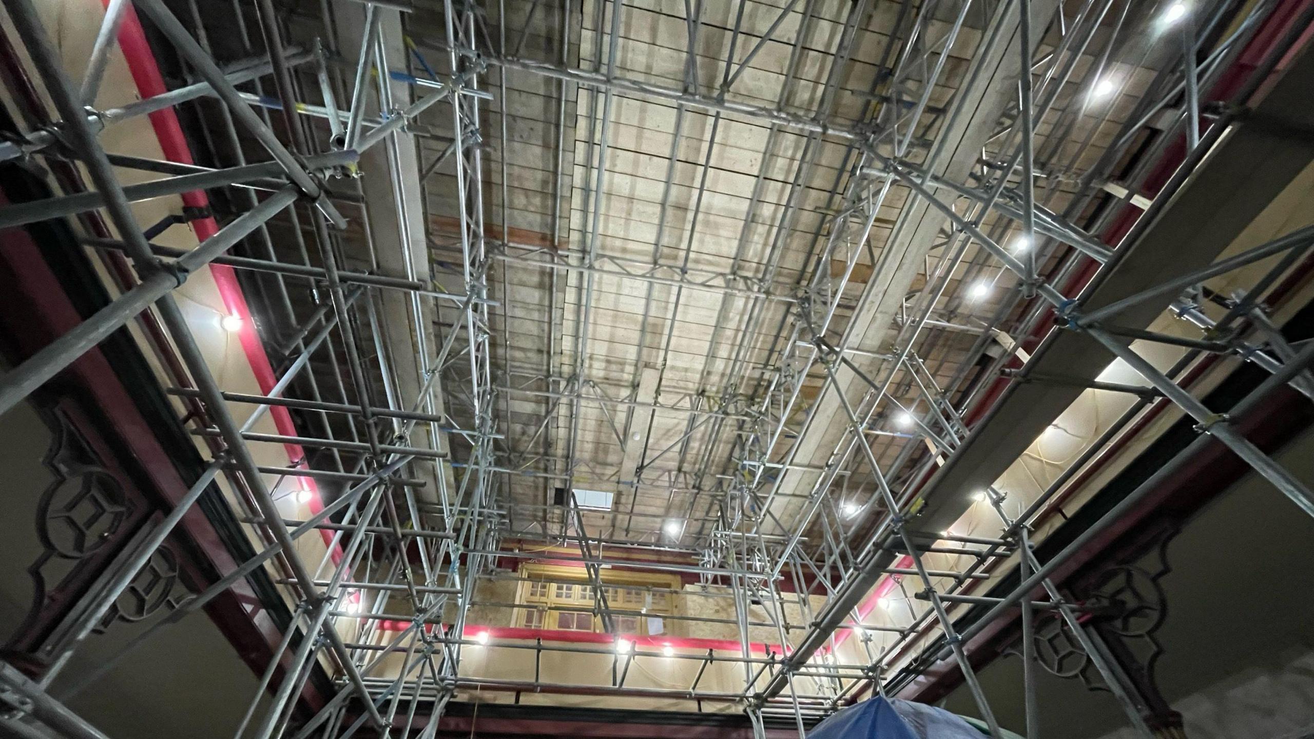 A photo from the Victorian Natural History Gallery. It is taken from a low vantage point and looks toward the room's ceiling. Huge amounts of scaffolding line the room. Lights can be seen placed along the scaffolding to illuminate room.