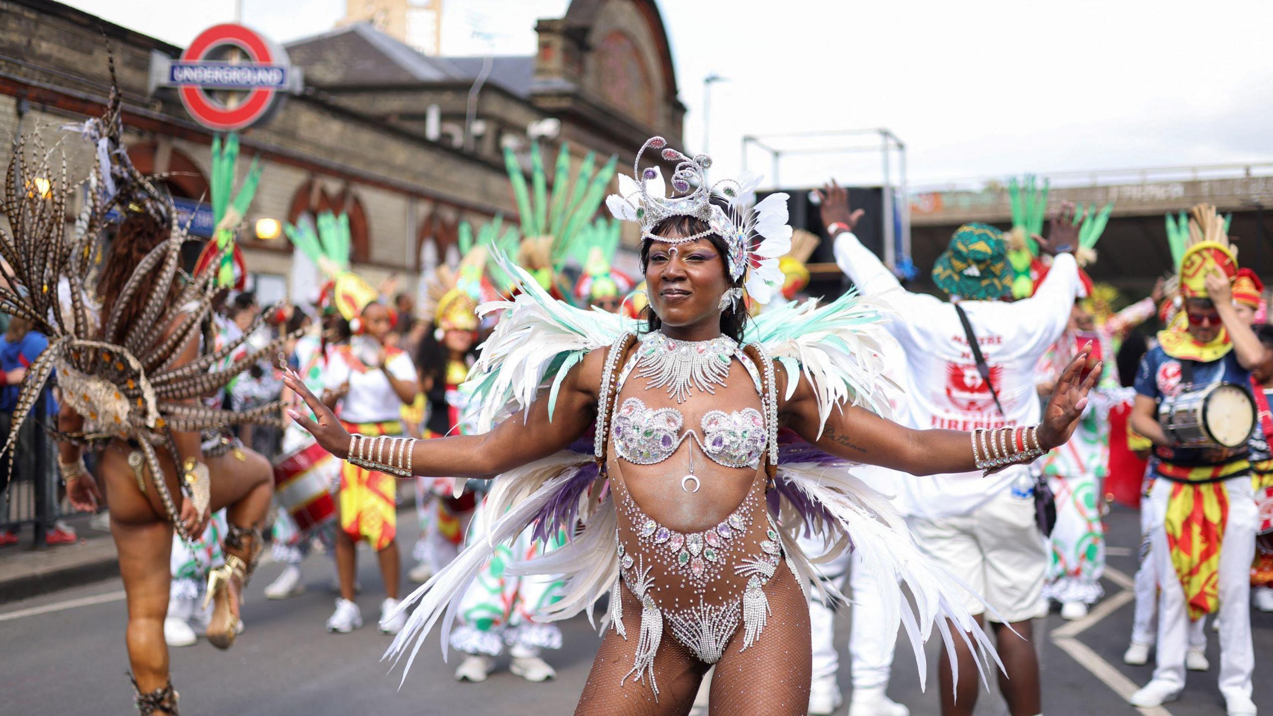 A black woman in a dazzling costume dances near a Tube station on the parade route