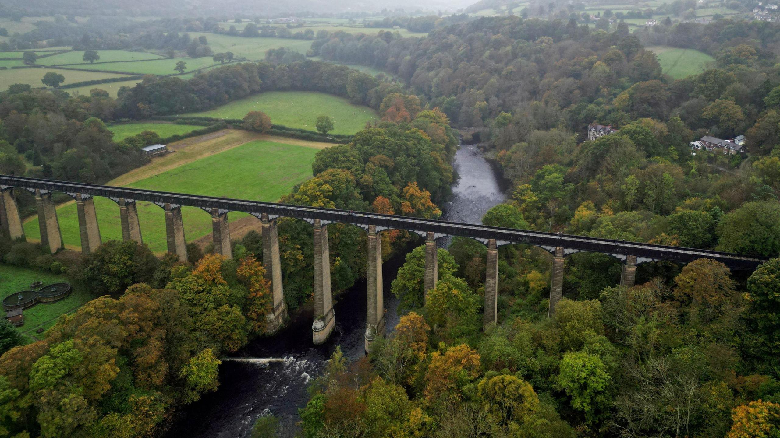 Arial shot of an arched aqueduct carrying a canal across a flad wide, wooded valley with a river running underneath it. The leaves of trees are turning yellow and brown and a coupe of houses are visible poking out of the woodland in the distance.