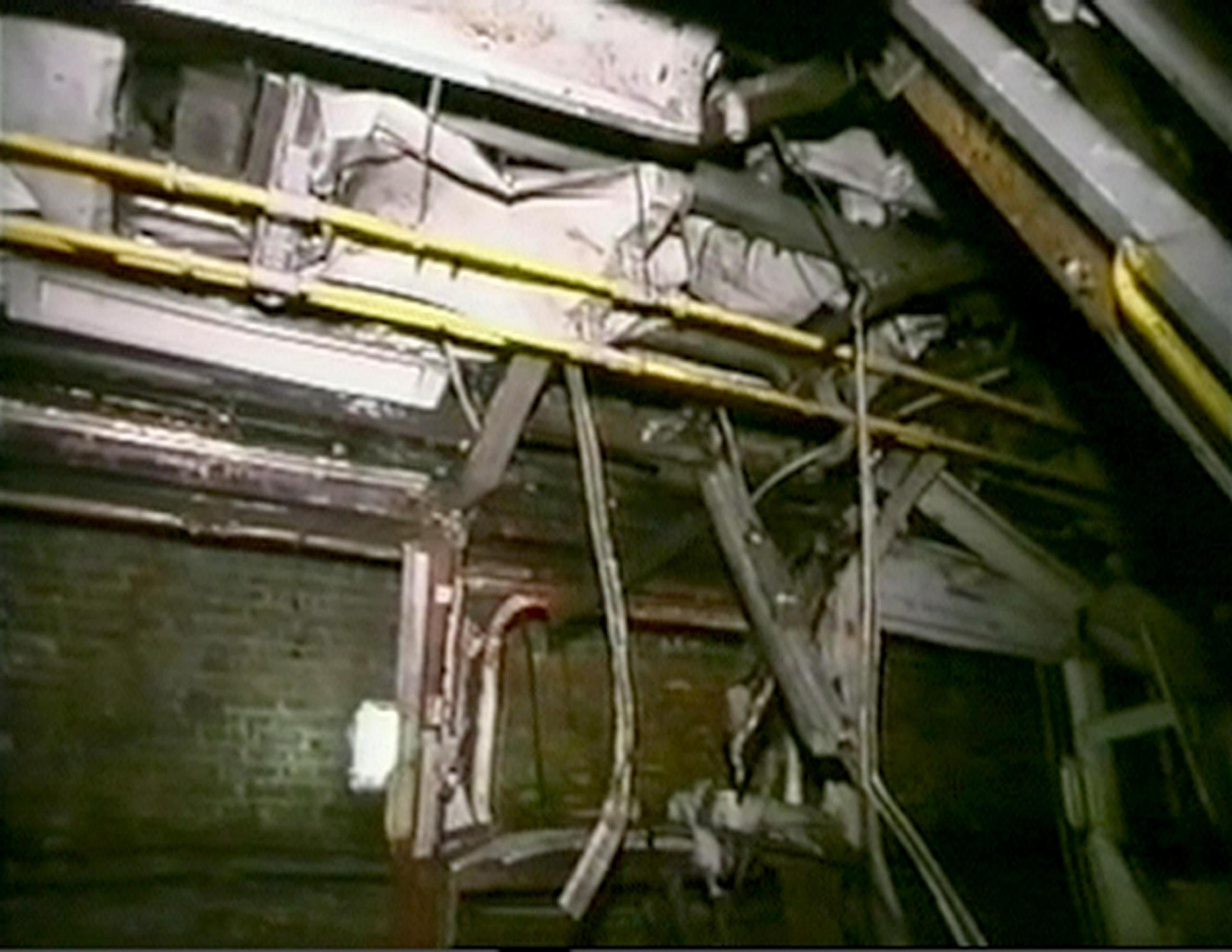 Screen grab taken from video footage taken by emergency services. The image shows the severely damaged ceiling of the Tube, with exposed wires and broken beams hanging down. Debris is scattered and the carriage no longer resembles a Tube carriage.