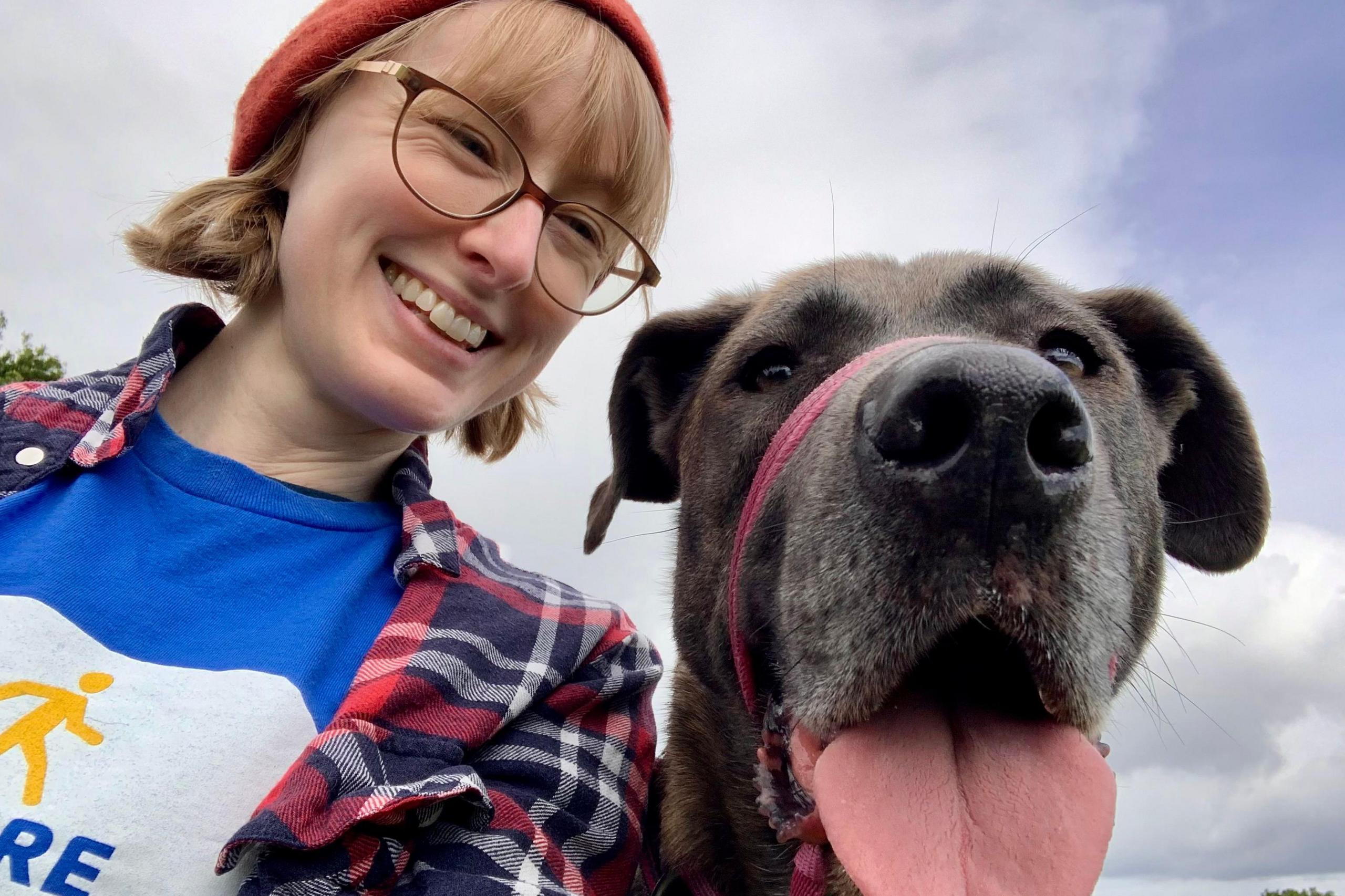 Alice Hildred with short blonde hair wearing an orange hat. She is smiling and is wearing glasses. She has a blue t-shirt, a chequered shirt and is with a brown dog which is sticking its tongue out.