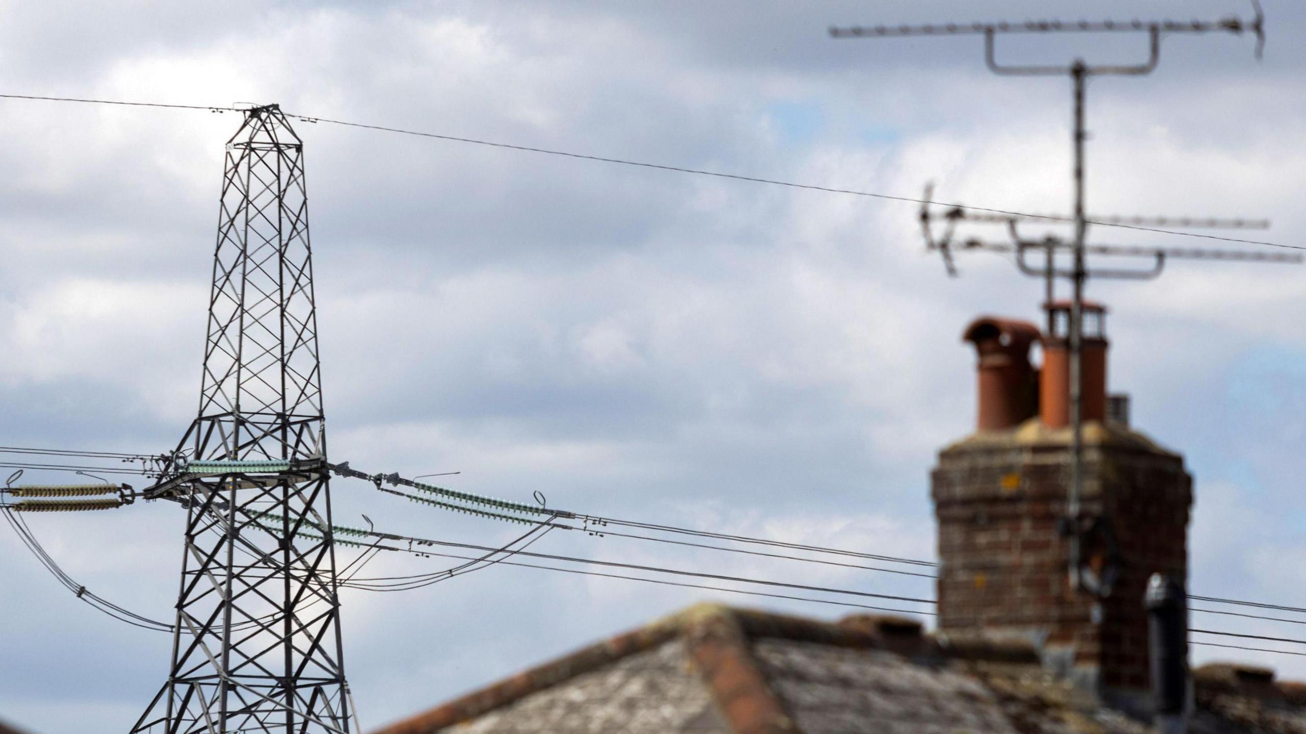 An electricity transmission tower and behind rooftops under a cloudy sky.