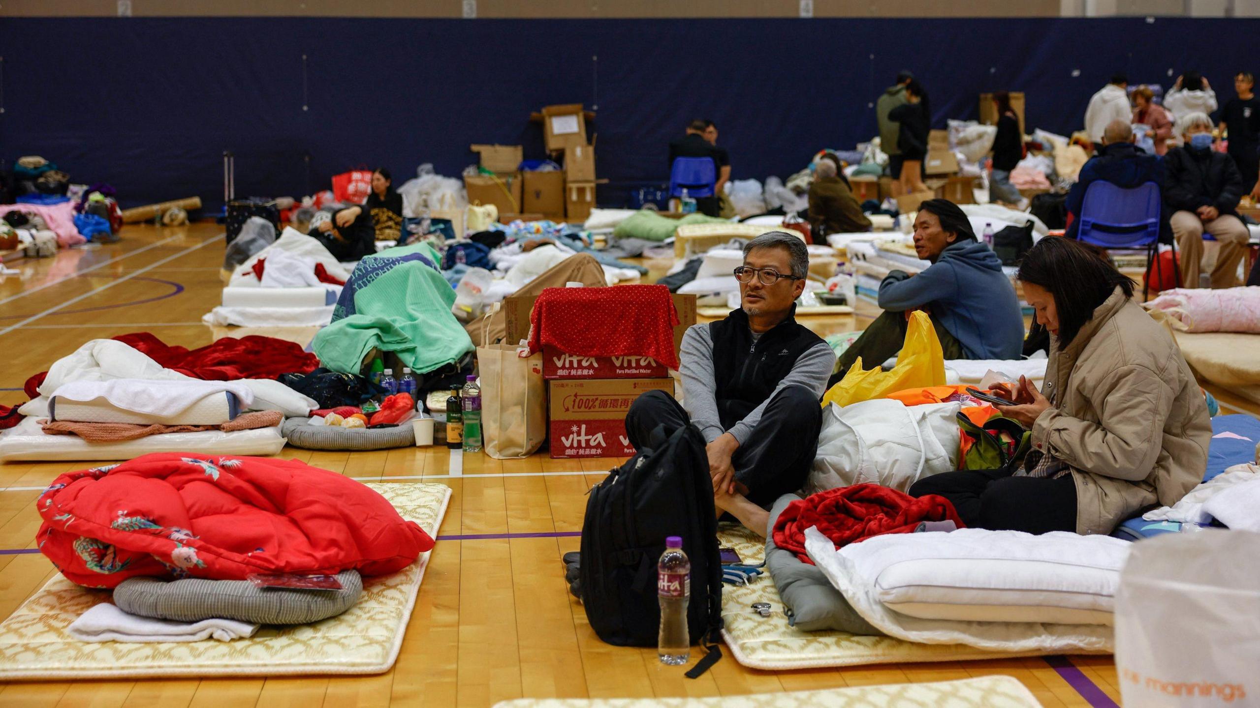 Mats have been laid on the floor for sleeping in a hall. There are people sitting and standing, and multiple boxes with supplies and people's personal belongings