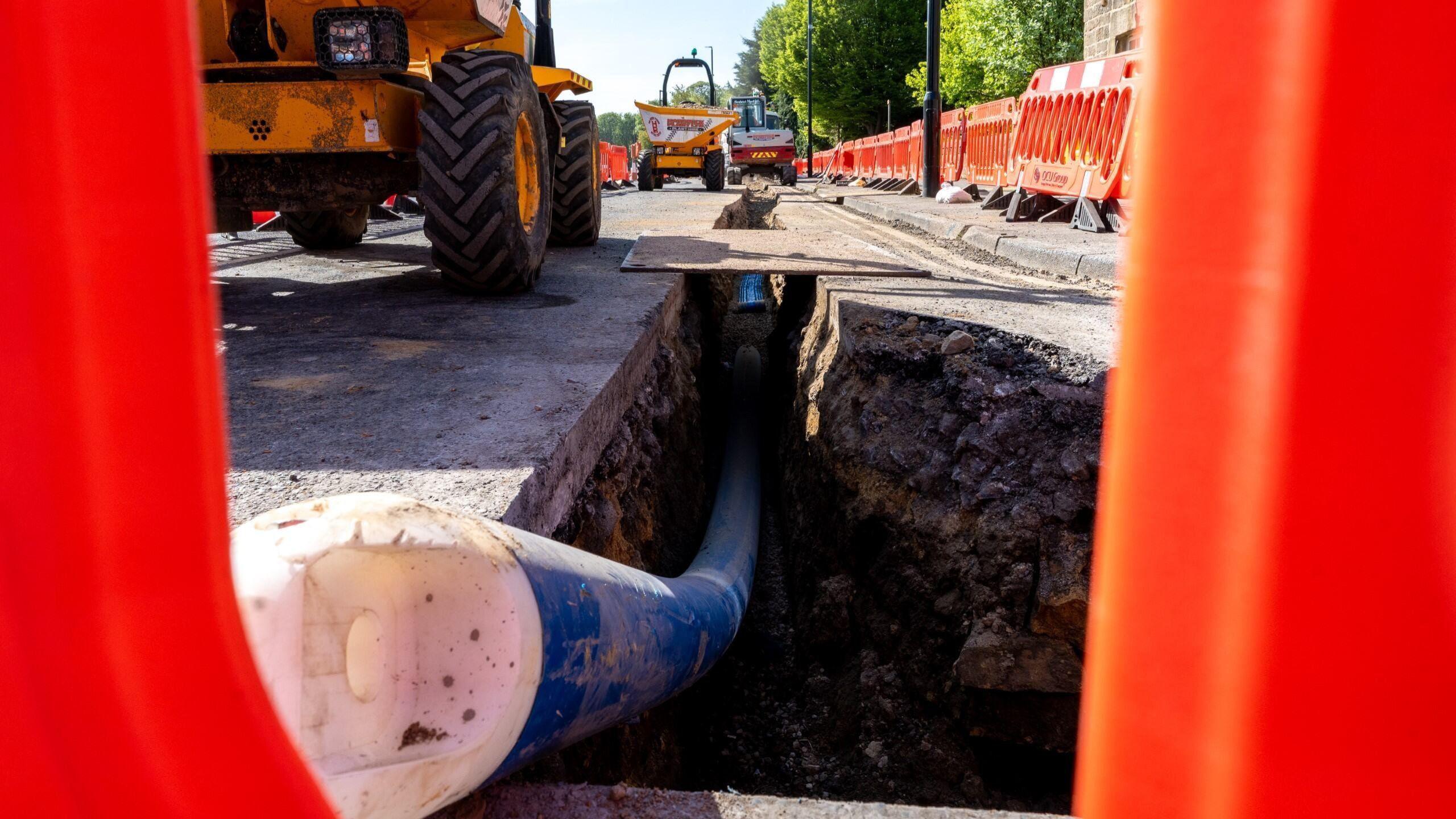 Construction vehicles sitting by a hole in the ground. Orange roadworks barriers line the street.