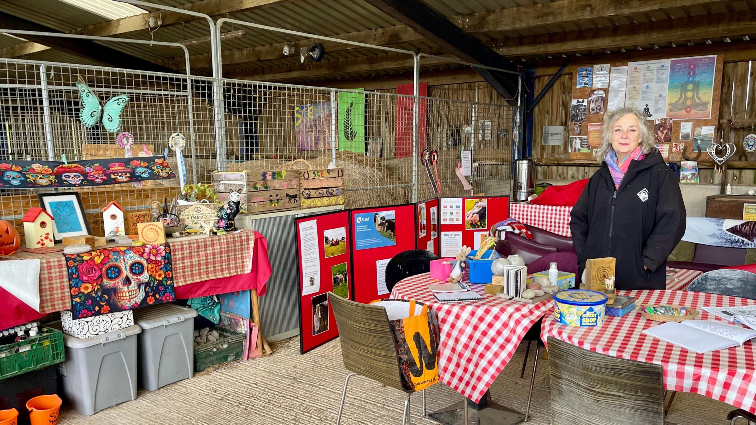A woman stands inside a stable which has been equipped like a classroom. There are tables, chairs, drawers, notcie boards and child friendly decorations. The woman is wearing a black coat, pink and purple scarf and has long grey hair. Haybales can be seen behind a steel grid.