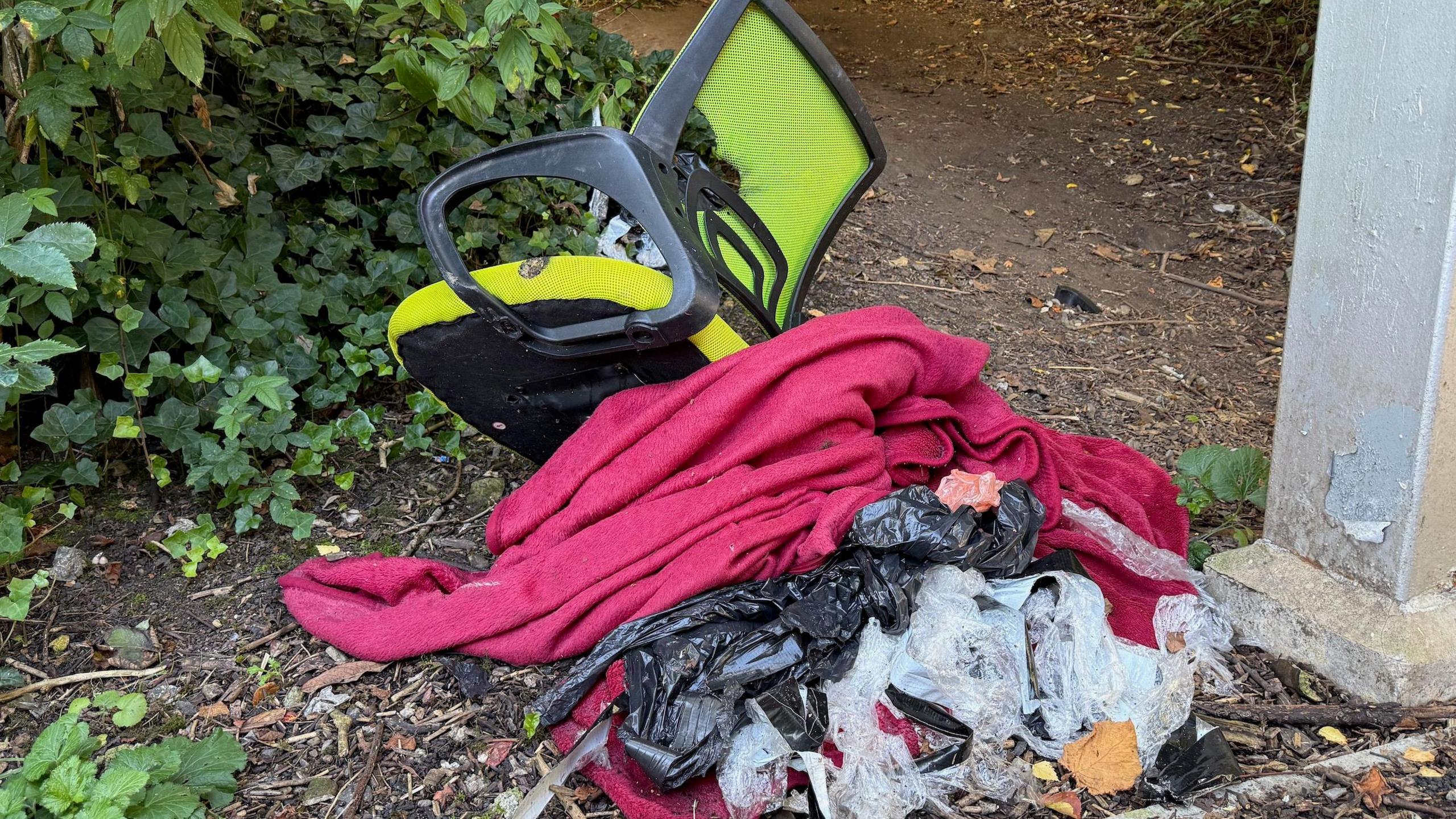 A bright green office chair dumped underneath a footbridge, along with an old red blanket and a bin bag, it's contents emptied across the ground