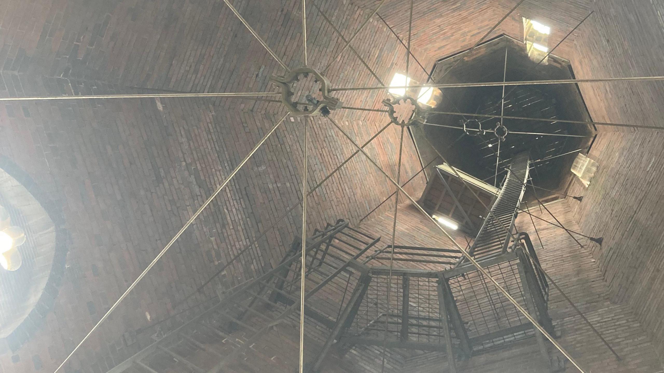 The picture is angled upwards, looking up into a church spire. It is brick and dimly lit. There are metal spokes in intervals all the way up, along with metal maintenance ladders and platforms. In a small window there is a wooden box for falcons to nest. Beyond that, there is a wooden grate at the top of the spire