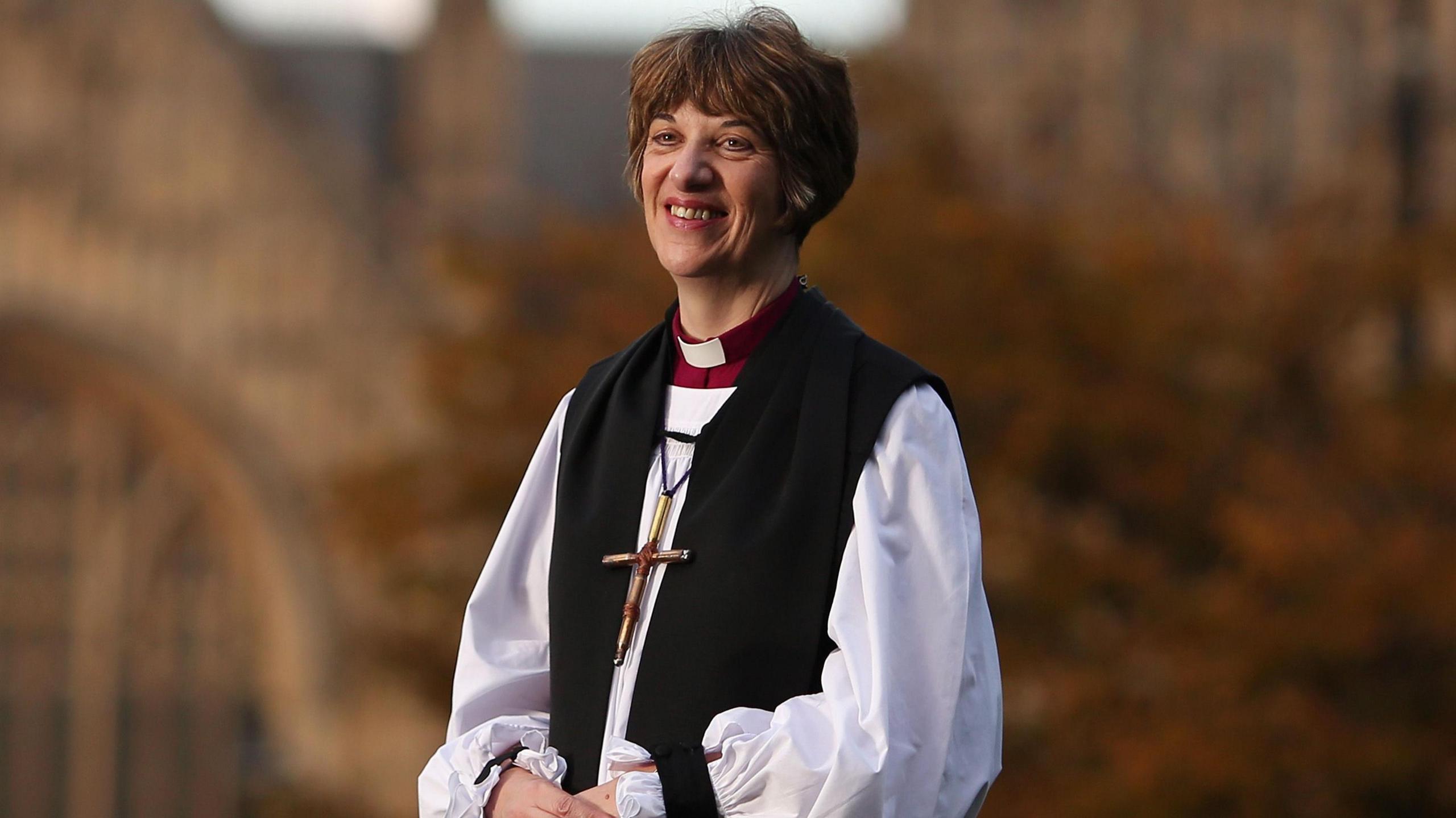 Bishop Treweek in her church garments, and wearing a large wooden crucifix, smiling in front of a brown backdrop