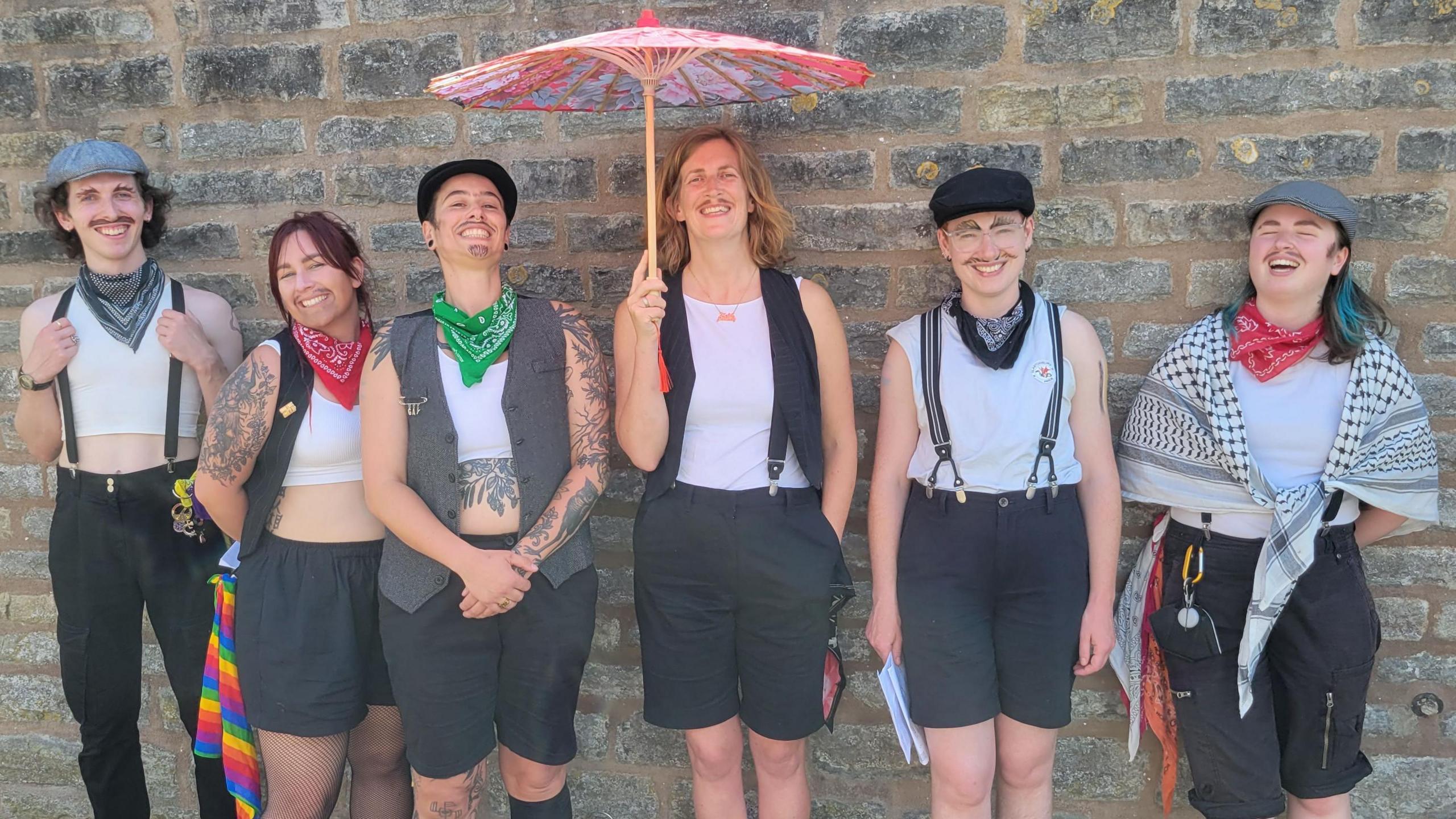 Six members of Molly No-Mates line up against a wall wearing white vest tops and black shorts, all with male-drag makeup. 