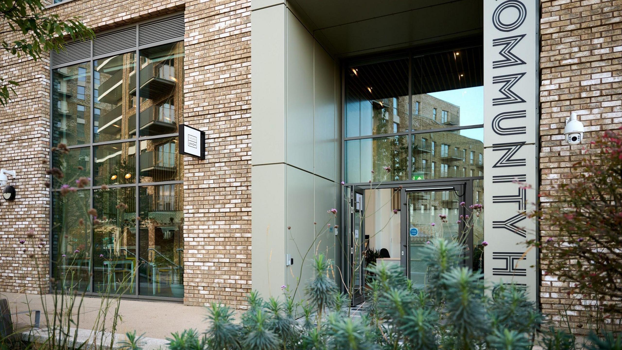 An external view of the hub - a brick and plaster building with big glass doors and windows and a large black and beige vertical sign reading 'community hub'. Green plants sit in the foreground