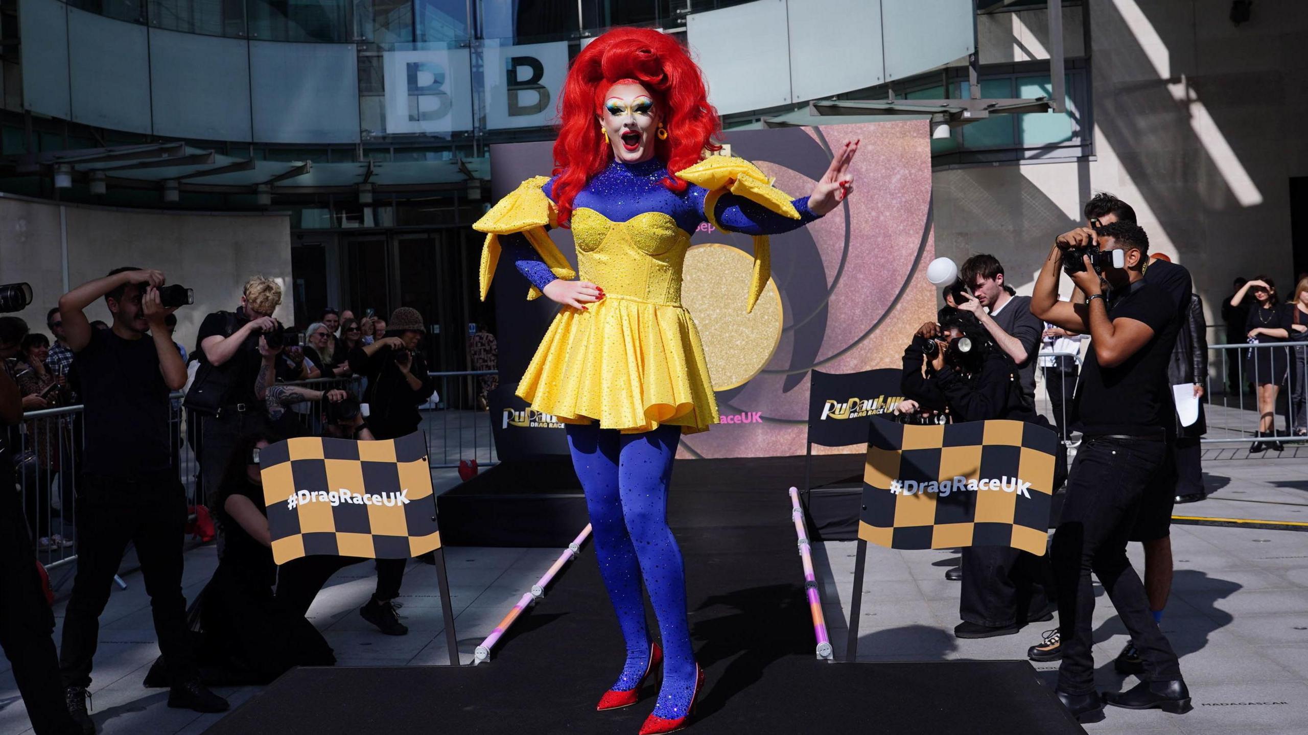 Drag queen Charra Tea wears a red wig, yellow bedazzled dress and long bedazzled blue sleeves. She is also wearing blue bedazzled tights and bright red heels. 