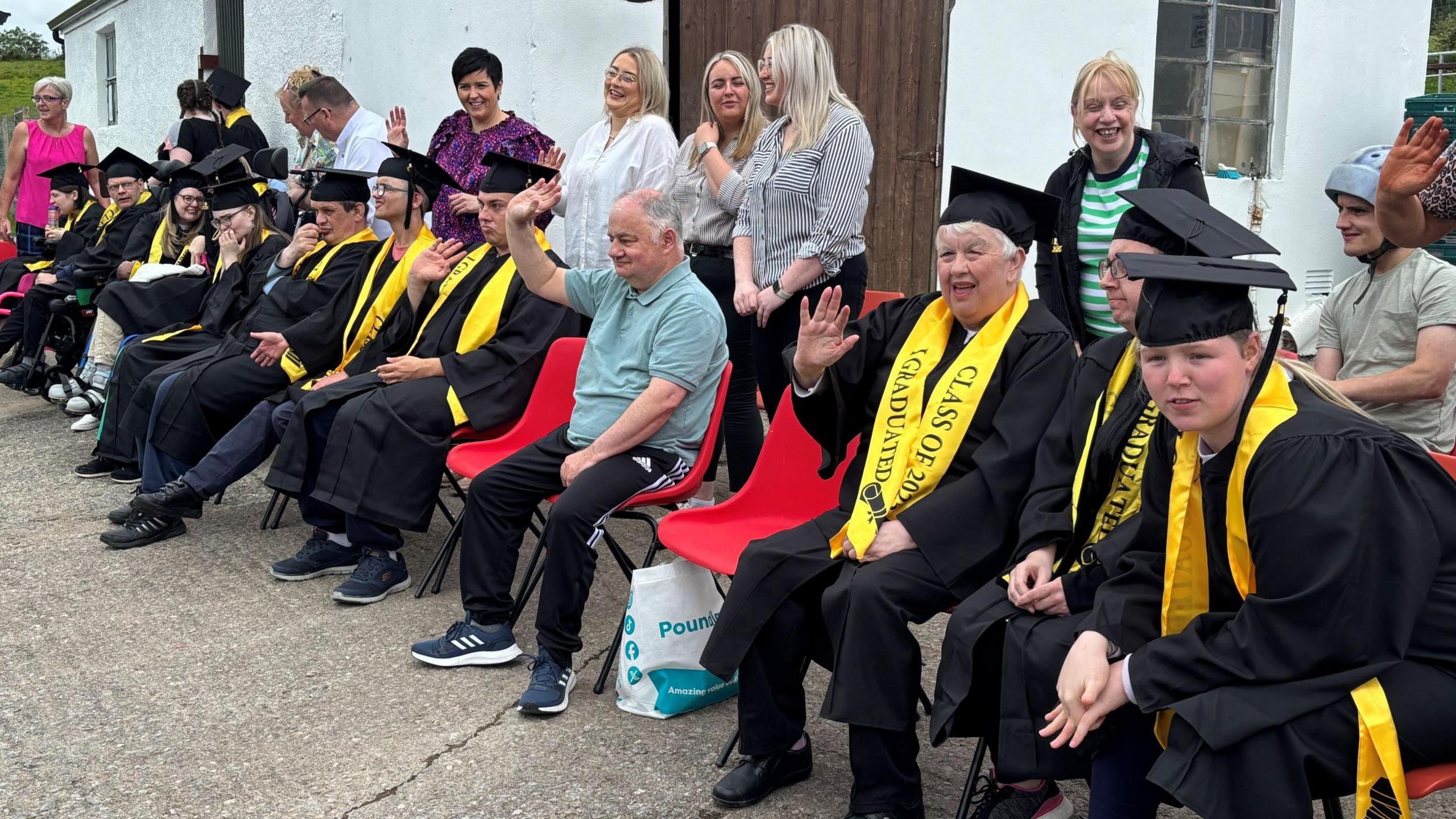 A row of people sitting on chairs. Most of them are wearing black graduation caps and gaps. They are also wearing yellow sashes which say "I graduated class of 2025".