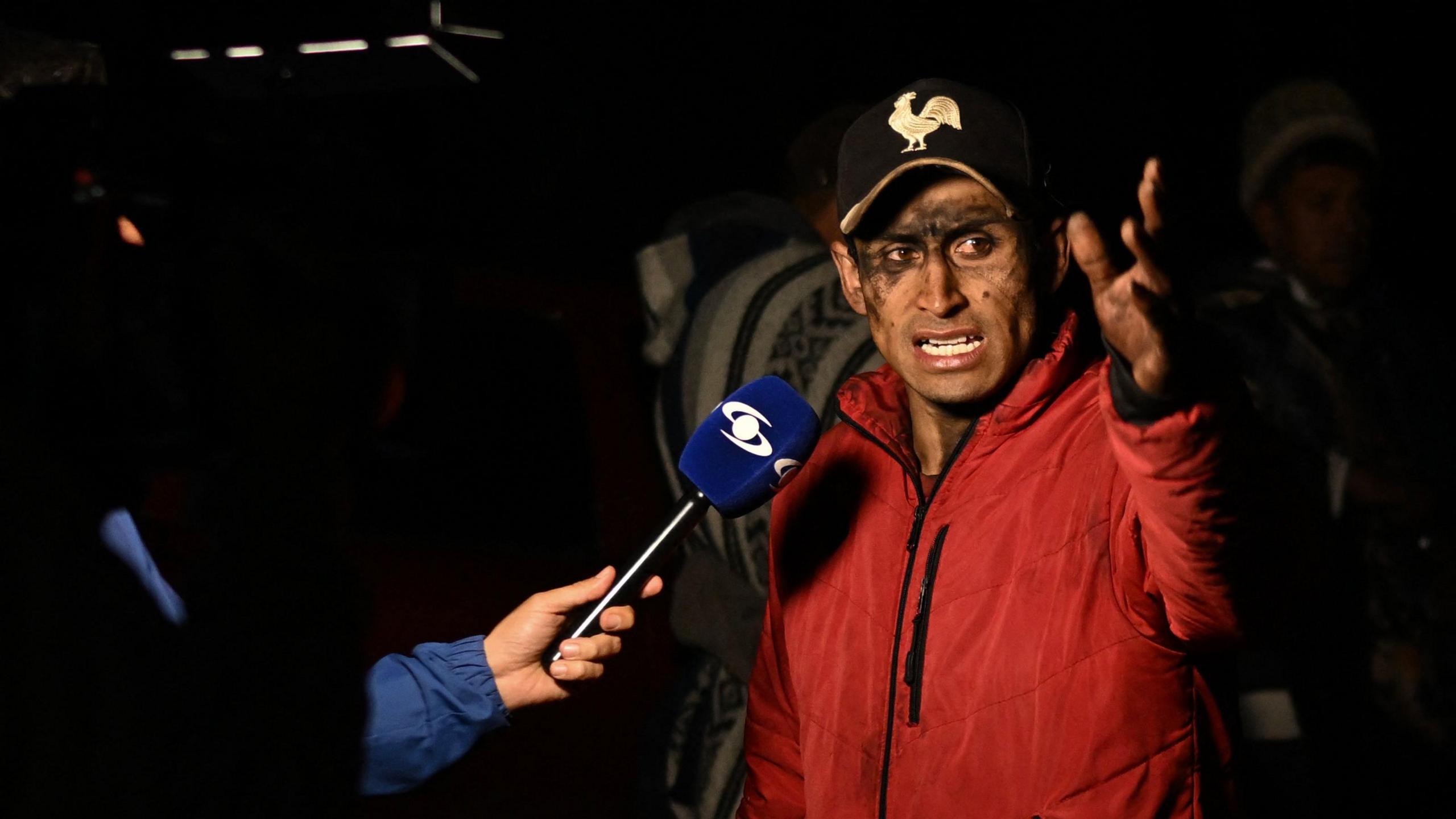 The relative of a miner caught in the explosion at the mine in Sutatausa gestures as he speaks to the media. His face is stained with coal dust. He is wearing a black baseball cap with a white cockerel embroidered on top and a red coat.