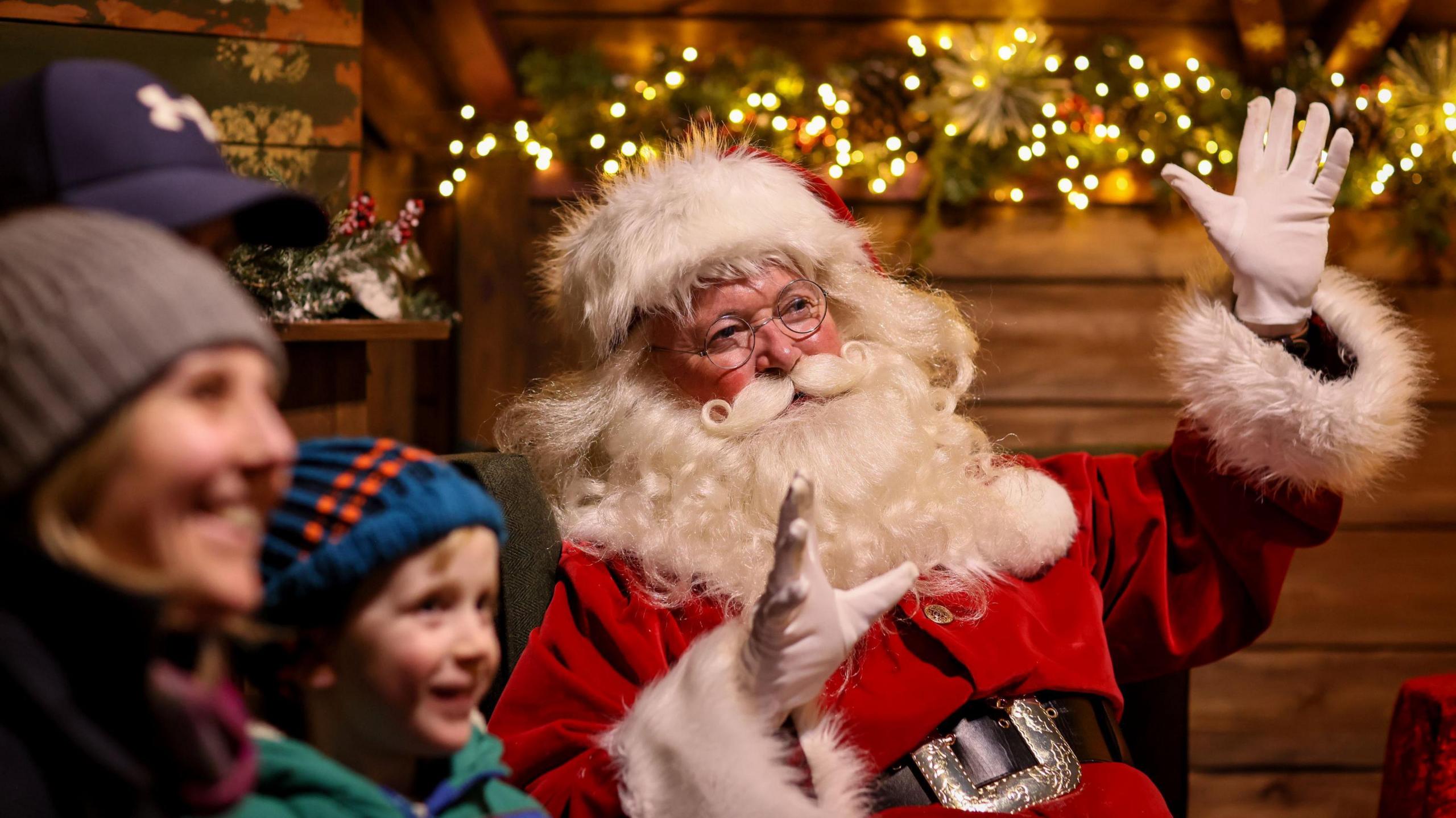 A man dressed as Father Christmas in a red outfit with white fur trim points to a display at Avon Valley Wildlife and Adventure Park, as a smiling family look in the same direction. Twinkling Christmas lights are visible in the background