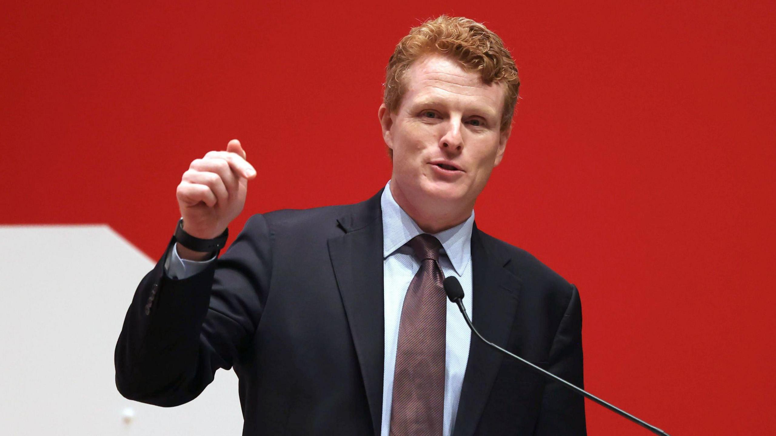 Joe Kennedy speaking a a microphone with his right hand raised. He is wearing a suit and tie, behind him a red and white background