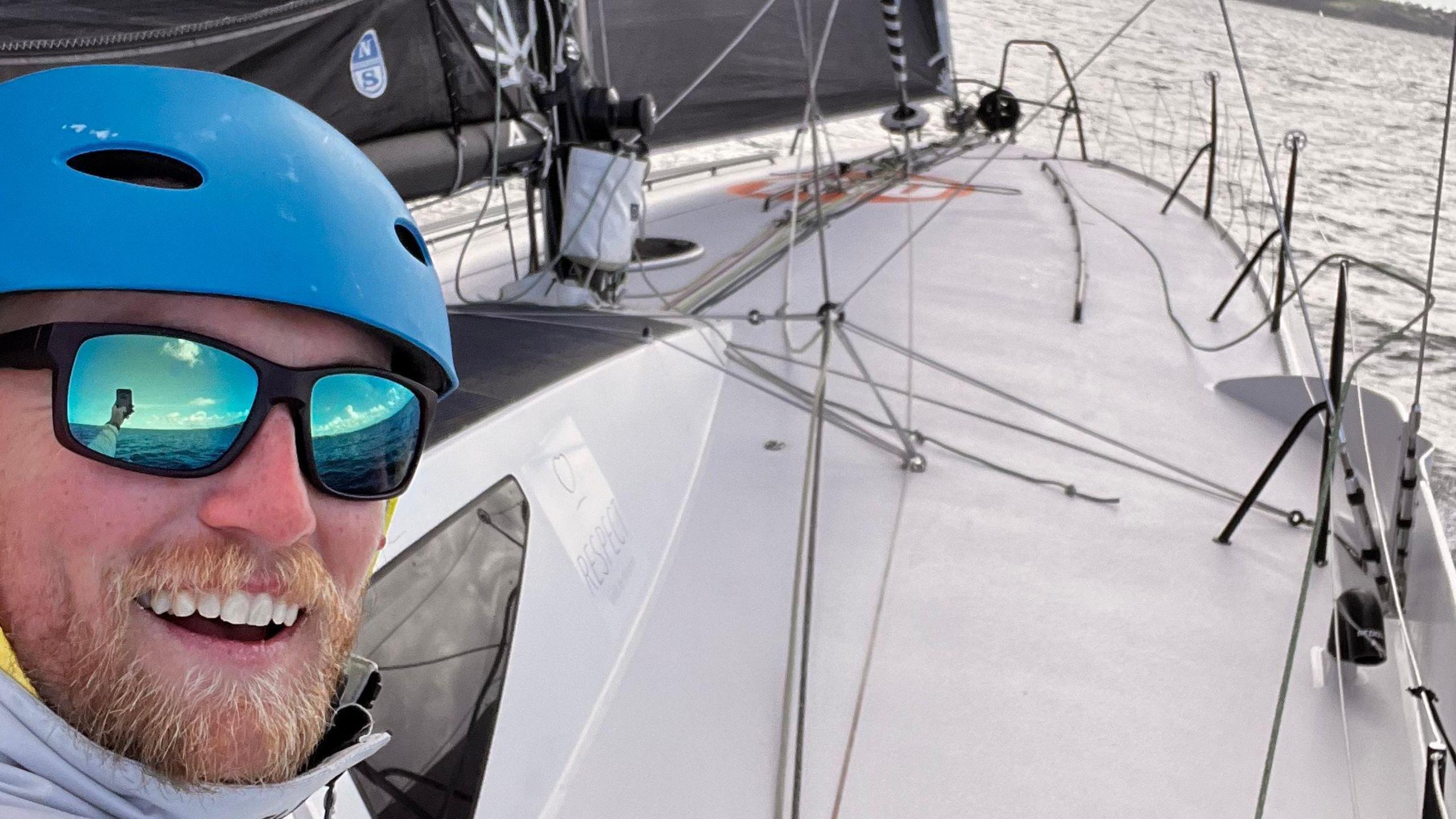 A picture of a man smiling at the camera while taking a selfie. He is wearing glasses and a helmet. The boat can be seen in the background.