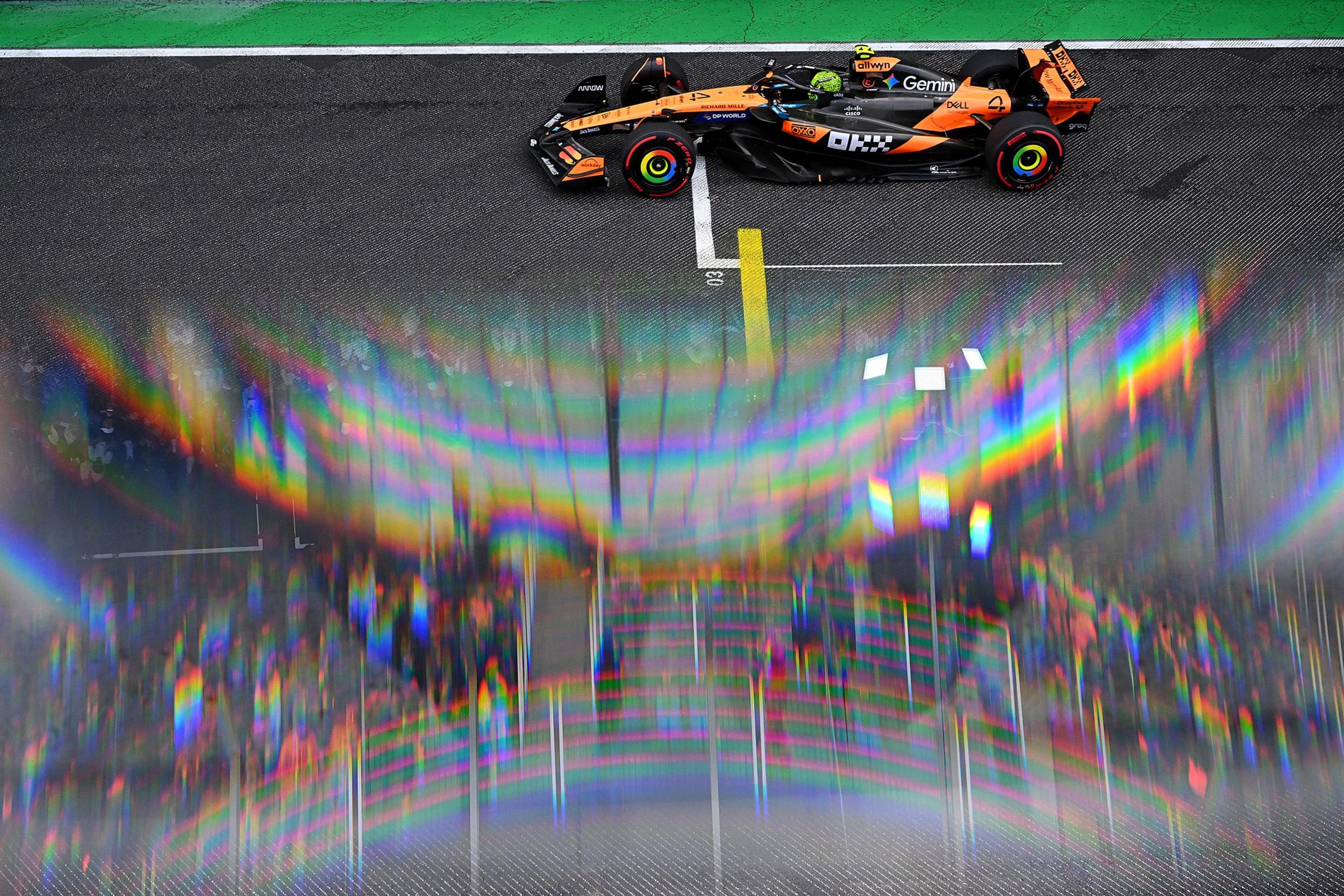 Orange and black Formula 1 car on a racetrack near the starting line, with green border visible at the top and vibrant rainbow-colored light reflections creating an abstract effect across the lower part of the image.