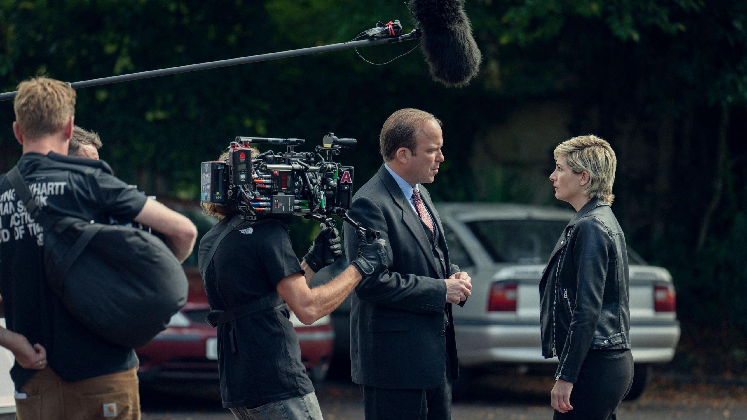 Rory Kinnear is speaking with Jodie Whittaker with a boom microphone above them and a man holding a camera stood nearby