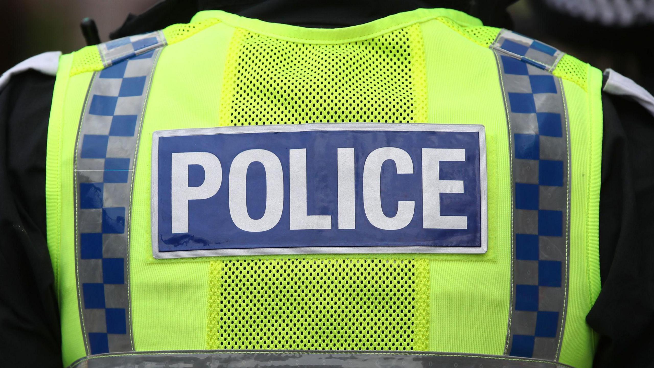 A police officer's upper back. Their vest is neon yellow with a blue label rectangular label with "police" written on it in white capital letters.