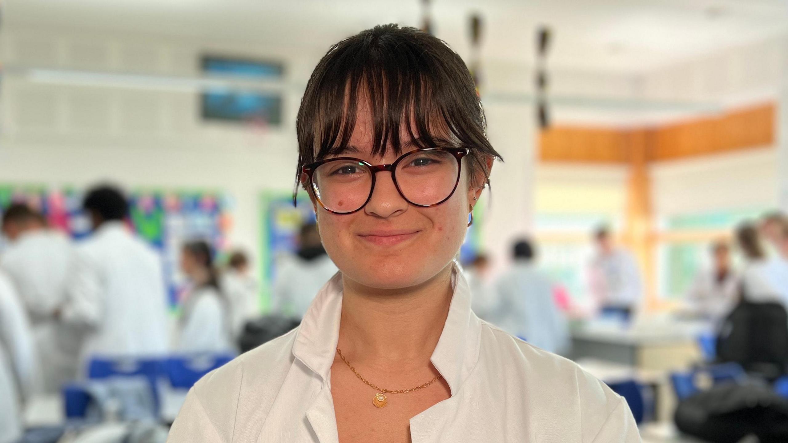 Johana is wearing a white lab coat stands in the foreground of a science classroom. The person's face is blurred for privacy. In the background, several other individuals, also in white lab coats, are engaged in various activities around laboratory tables. The room is well-lit with large windows on one side and colorful decorations on the walls. There are blue chairs and desks scattered throughout the room.