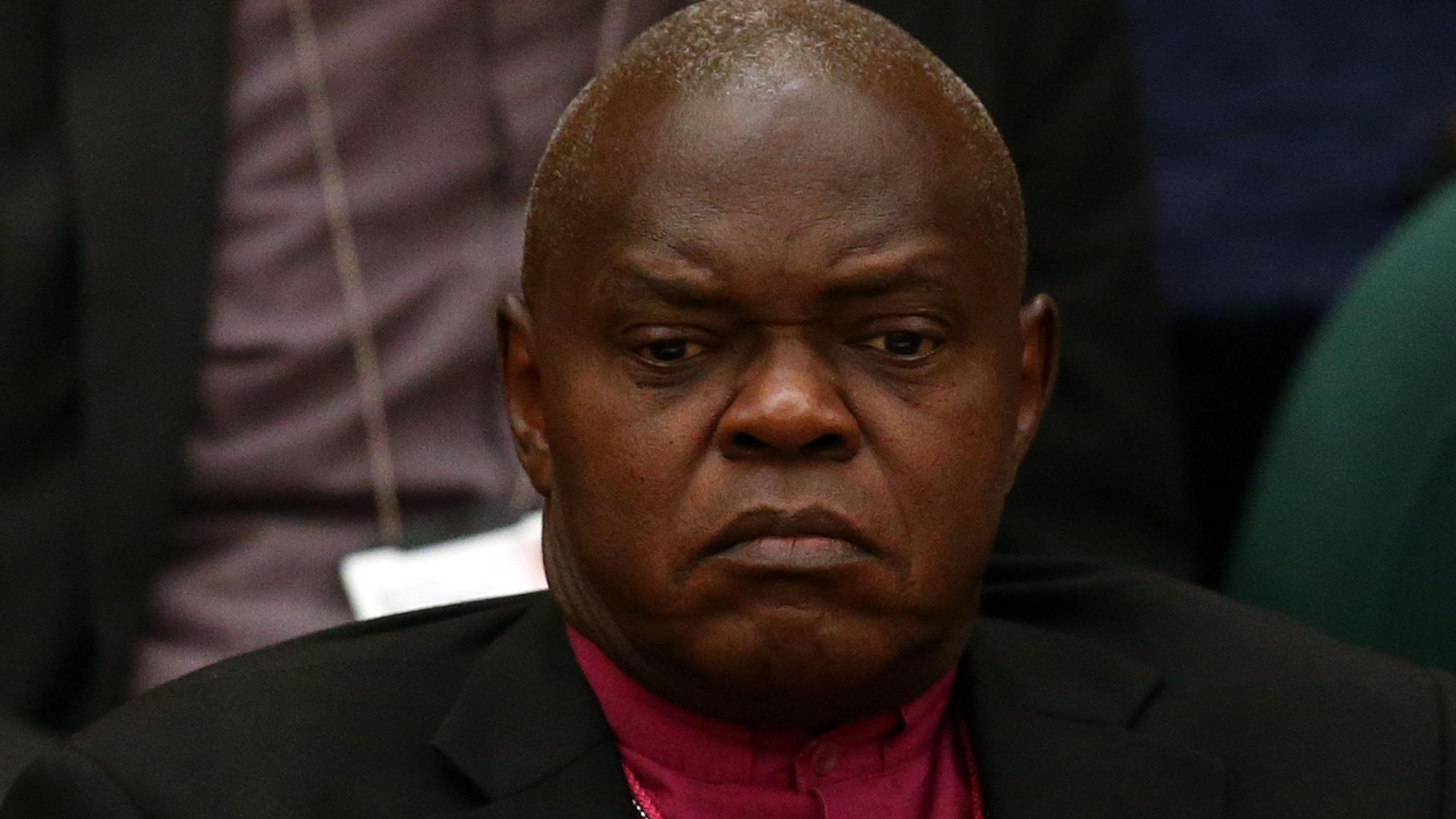 Lord Sentamu, formerly the Archbishop of York, dressed in a black jacket and purple vestments. He is gazing, apparently absent-mindedly, to somewhere beneath the camera.