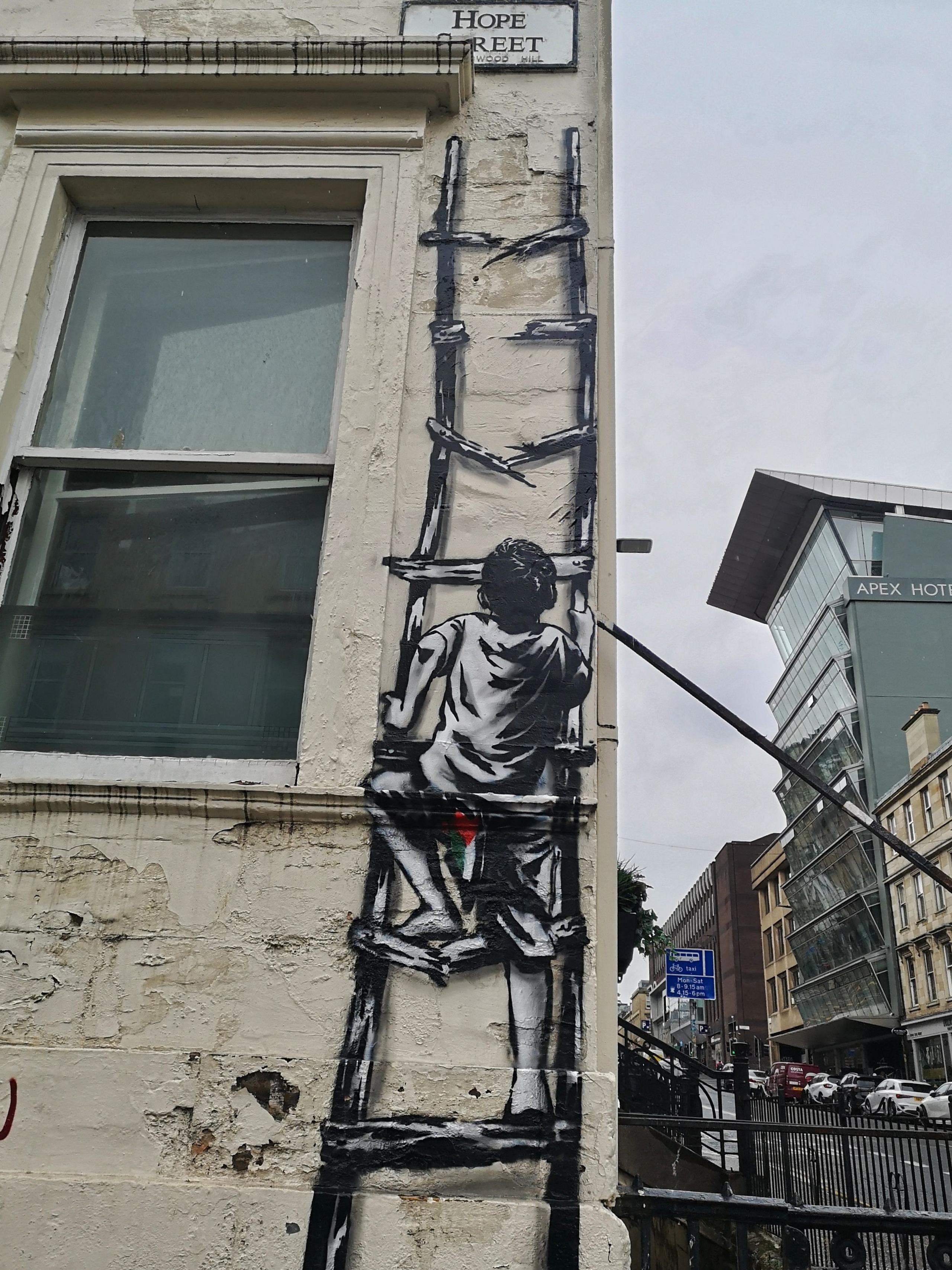 A street art mural painted on the side of a building features a person climbing a ladder, which is part of the mural itself, giving the illusion of ascent. The figure is viewed from behind, appearing to climb toward the top of the building. Above the mural, there's a street sign that reads "Hope Street".