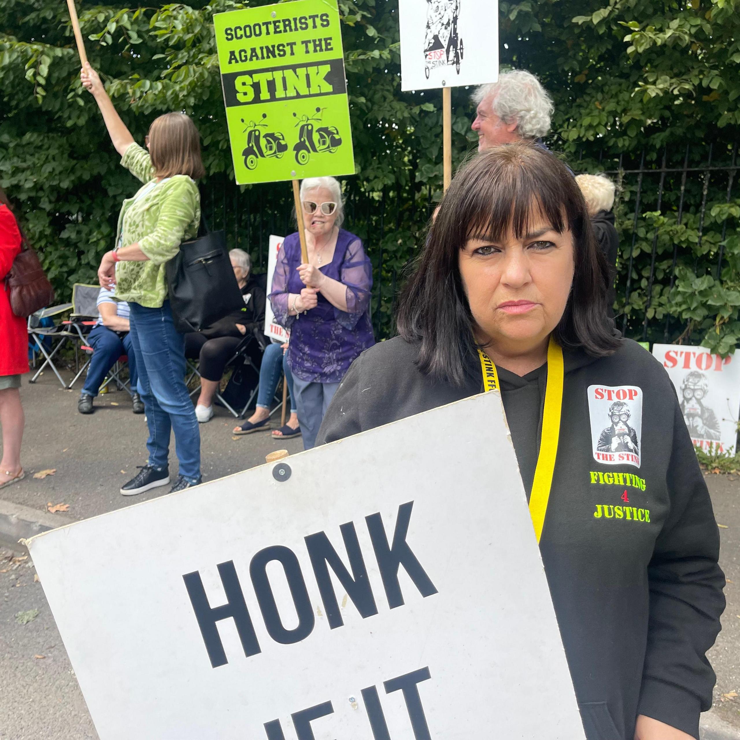 A woman with long black hair wearing a black top that says "stop the stink" and "fighting for justice" and holding a white sign that says "honk if it stinks".