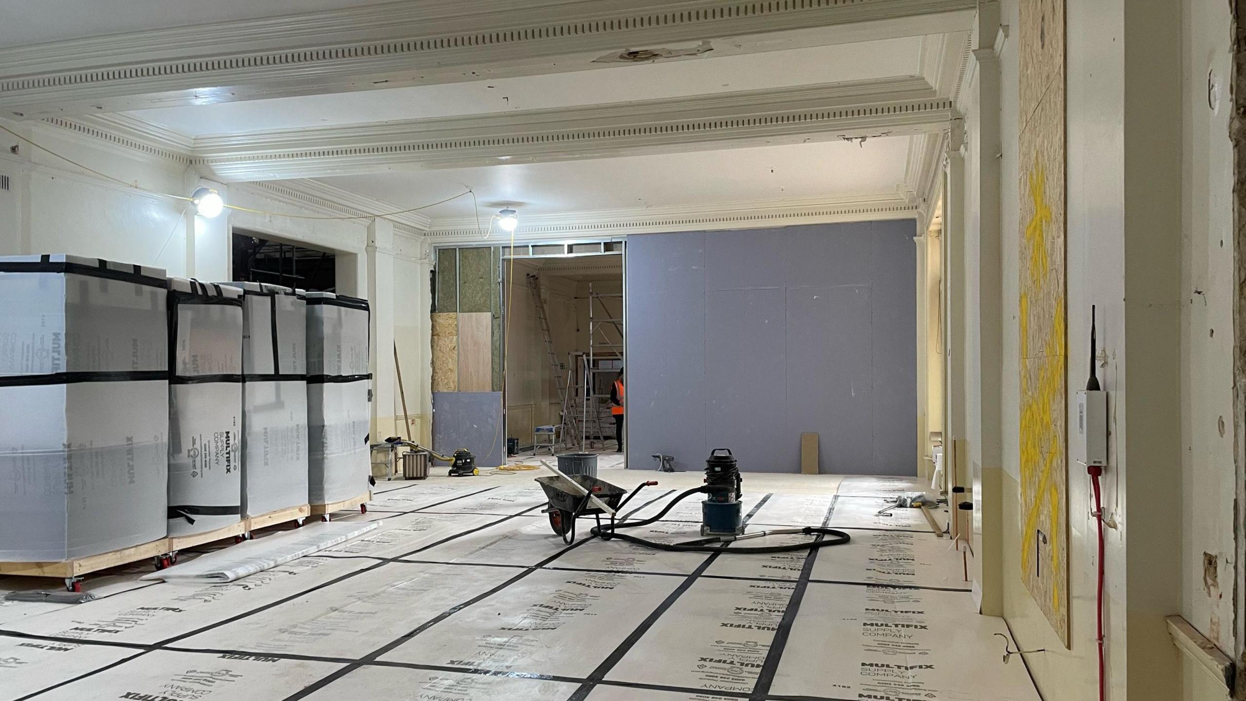 A room in the museum is pictured. Cardboard sheets have been placed on the flood and there is construction items dotted about the room. Large boxes have been placed on one site of the room.