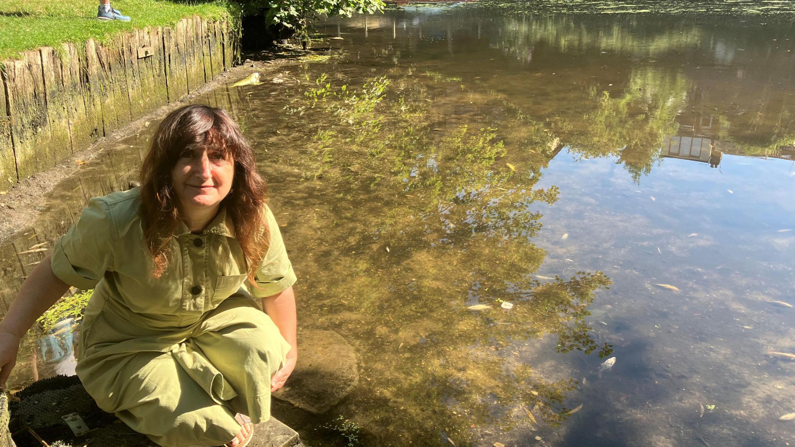 A woman wearing a light green jumpsuit kneeling down by the side of the water. 
She has wavy brown hair and is smiling while looking up to the camera. She is kneeling next to the river.