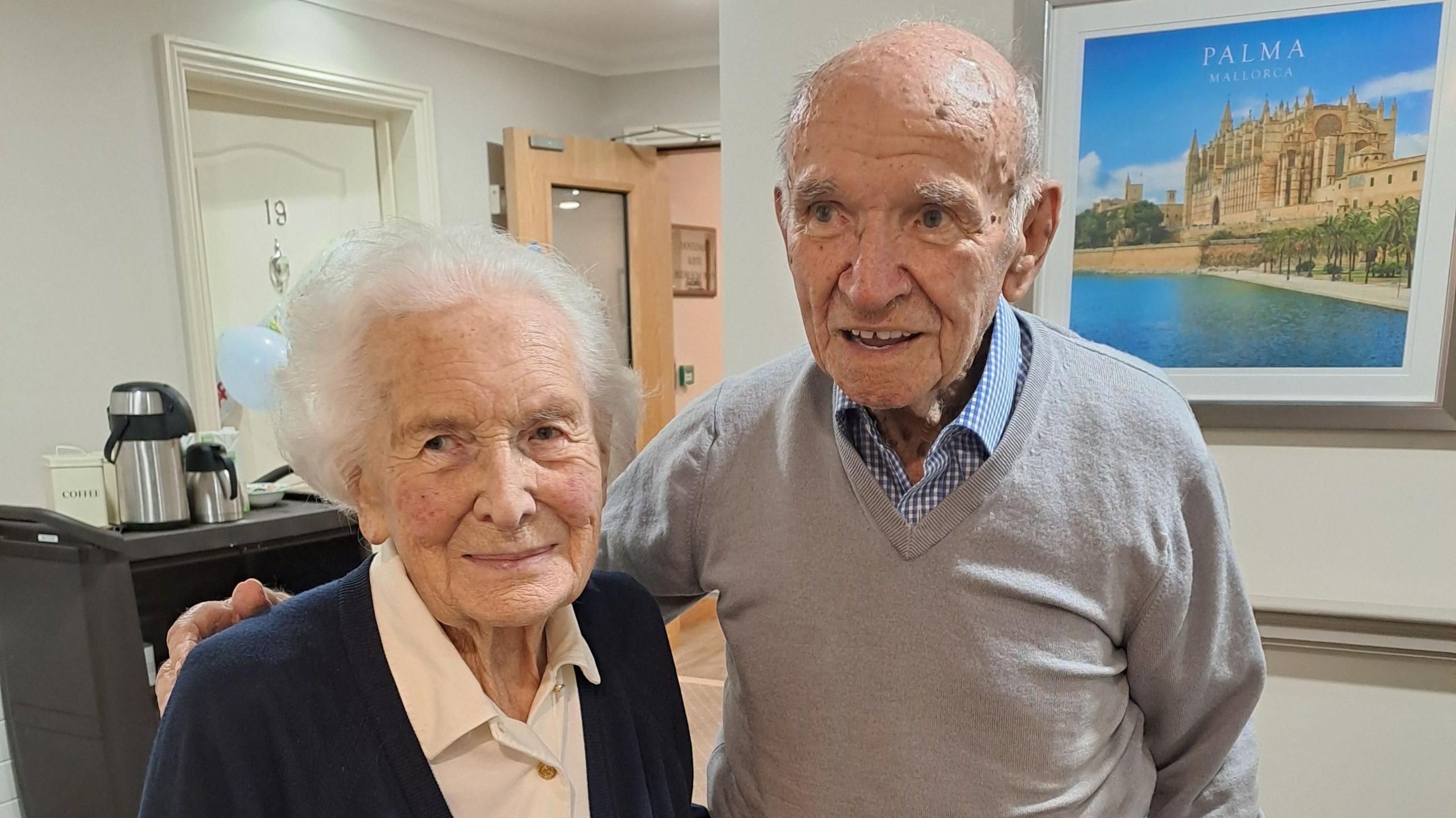 Trudy and Alan stand together. Alan has his arm around Trudy, she wears a navy cardigan over a cream blouse and he wears a grey jumper over a blue checked shirt.