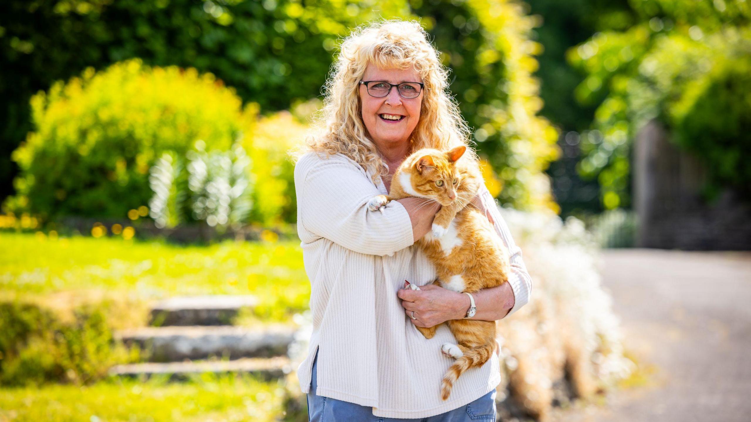 Photograph of Briony and Fizz, bundled up in Briony's arms. Green gardens can be seen behind them as the sun shines in the background. Briony has blonde curly hair and smiles at the camera. 