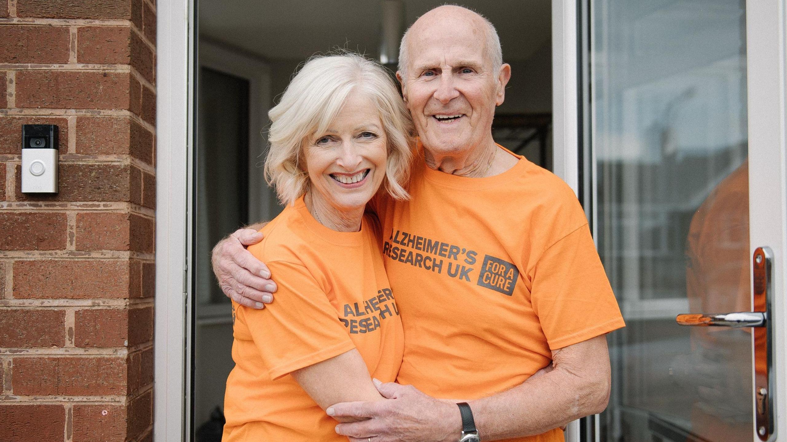 A man and woman, wearing orange t-shirts with "Alzheimer's Research UK - For a Cure" written on the front, embrace in front of an open door.