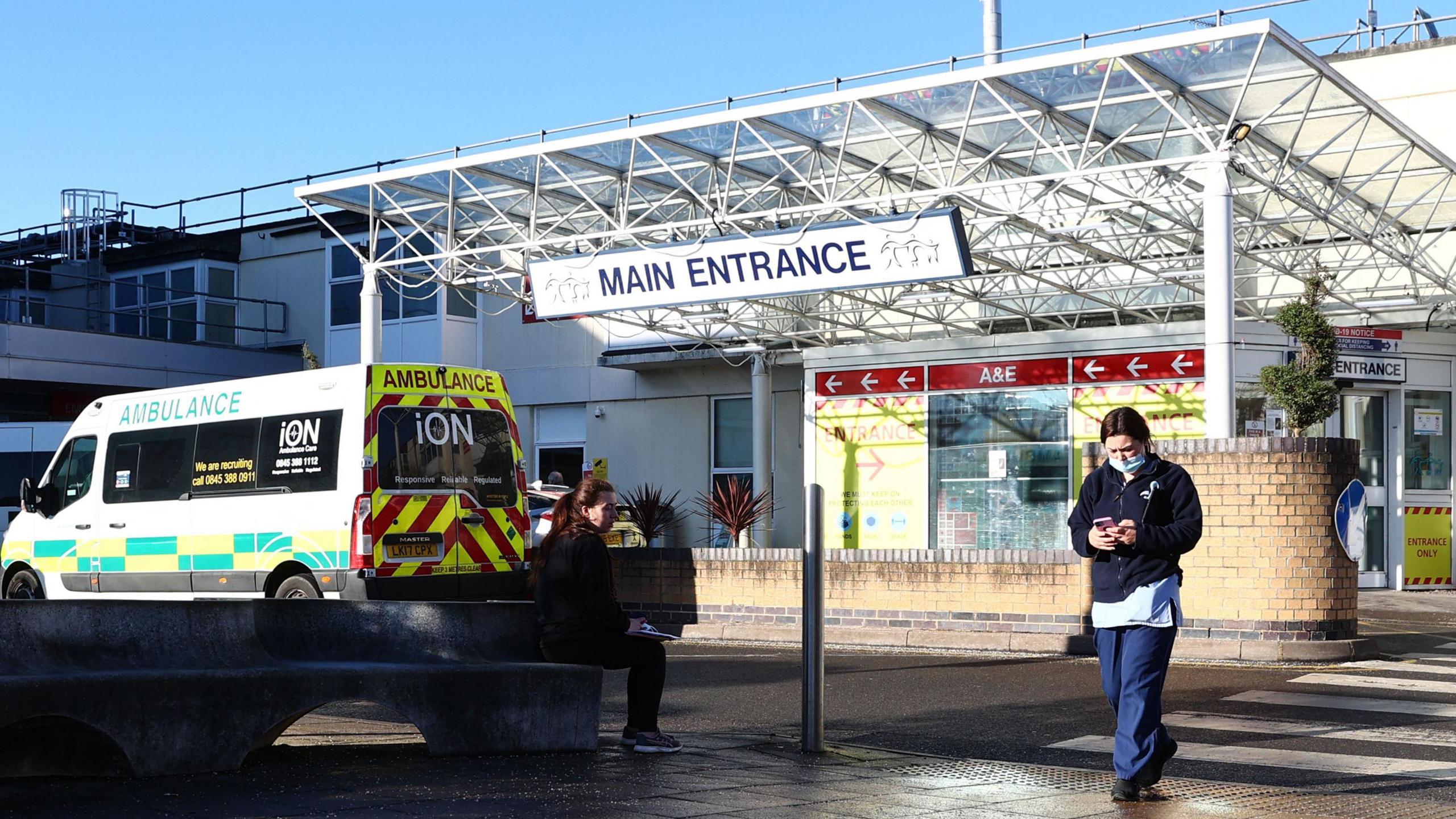 The main entrance of a hospital. An ambulance is parked outside. A person walks towards the camera across a zebra crossing